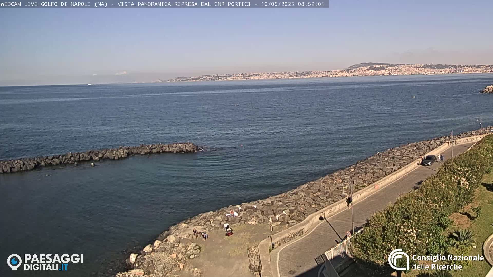 A rocky shoreline with a paved road and a few people is visible, overlooking a calm sea and a distant city under a clear sky.