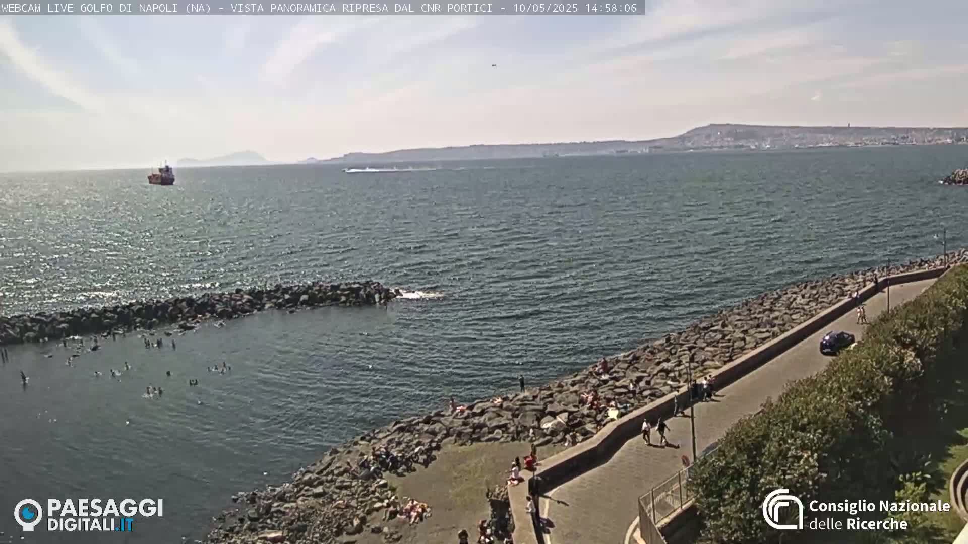 A partly sunny day shows a rocky shoreline with people sunbathing and swimming in a calm sea, a distant ship, and a city in the background.