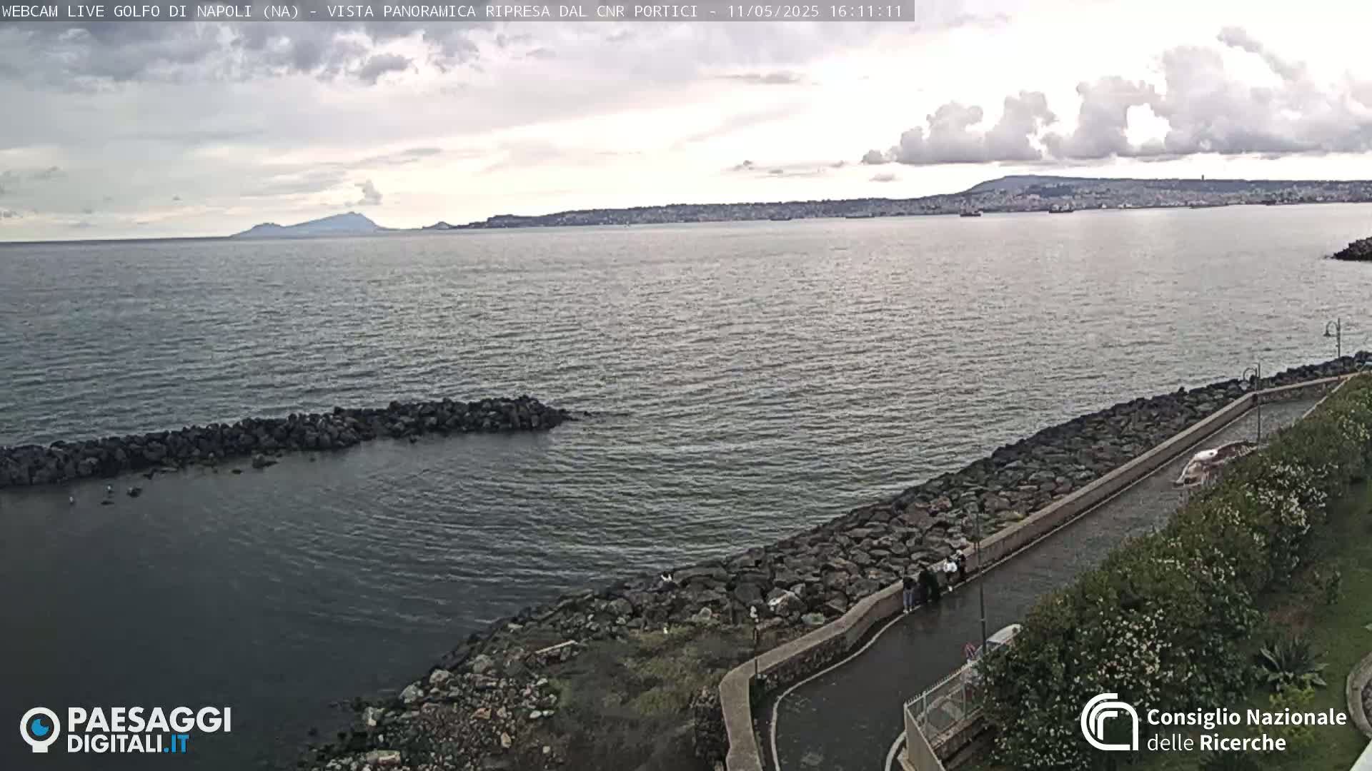 A calm, overcast day shows a rocky shoreline and body of water, with a distant mountain range visible across the bay.