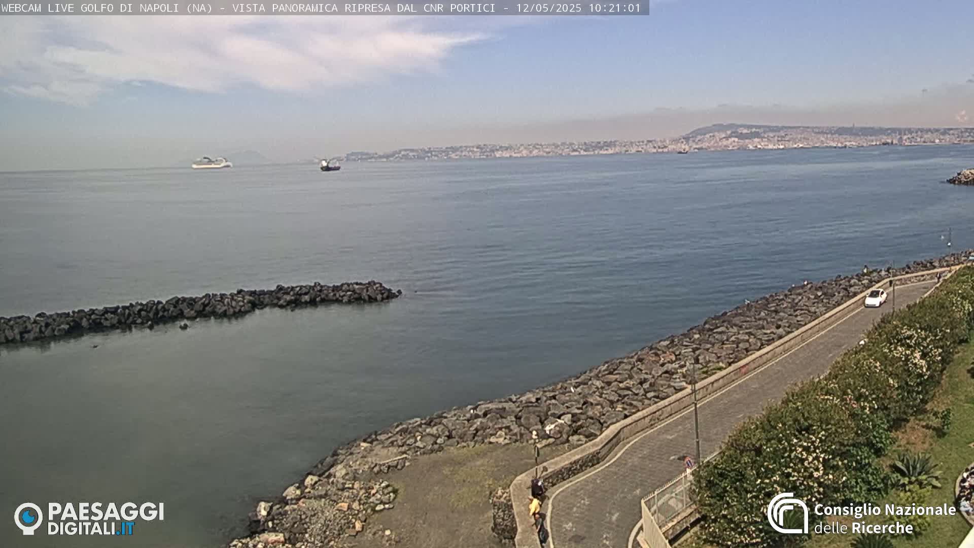 A coastal road curves along a rocky shoreline beside a calm bay, with a city and distant mountains visible across the water under a mostly clear sky.