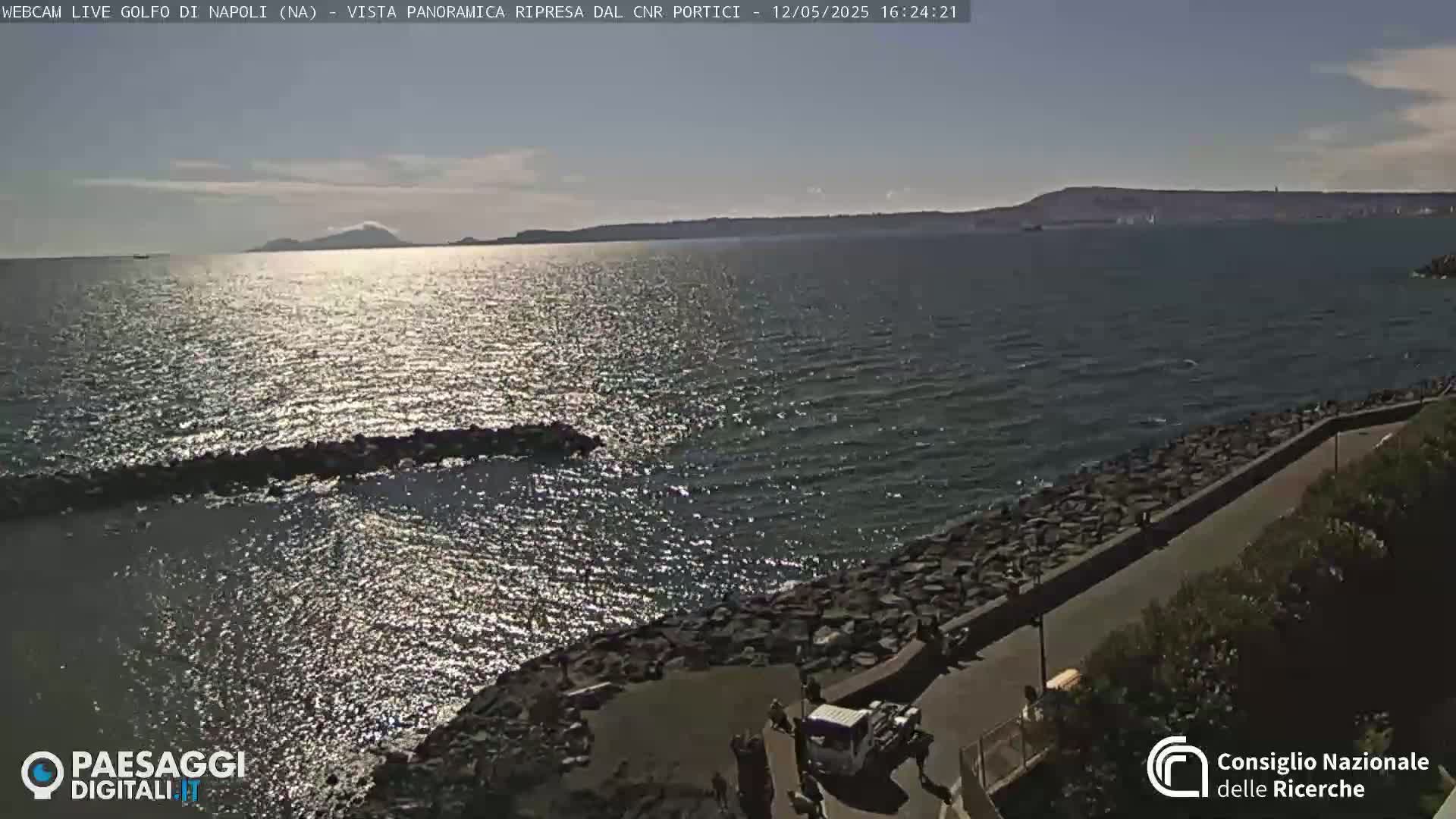 A rocky shoreline with a paved path overlooks a sunlit sea, distant mountains visible on the horizon under a mostly clear sky.