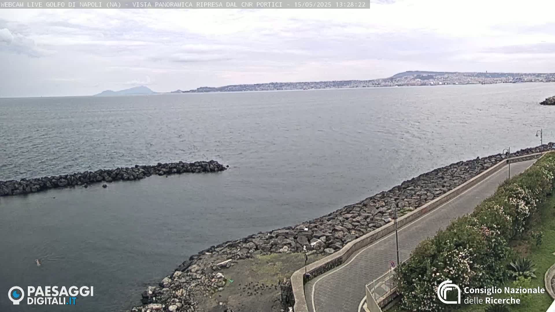 A rocky shoreline curves gently alongside a paved road, overlooking a calm sea with a distant, hazy mountain range under an overcast sky.