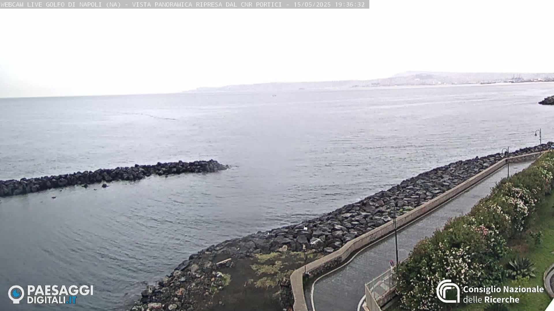 A rocky coastline with a calm sea stretches toward a distant city under an overcast sky.