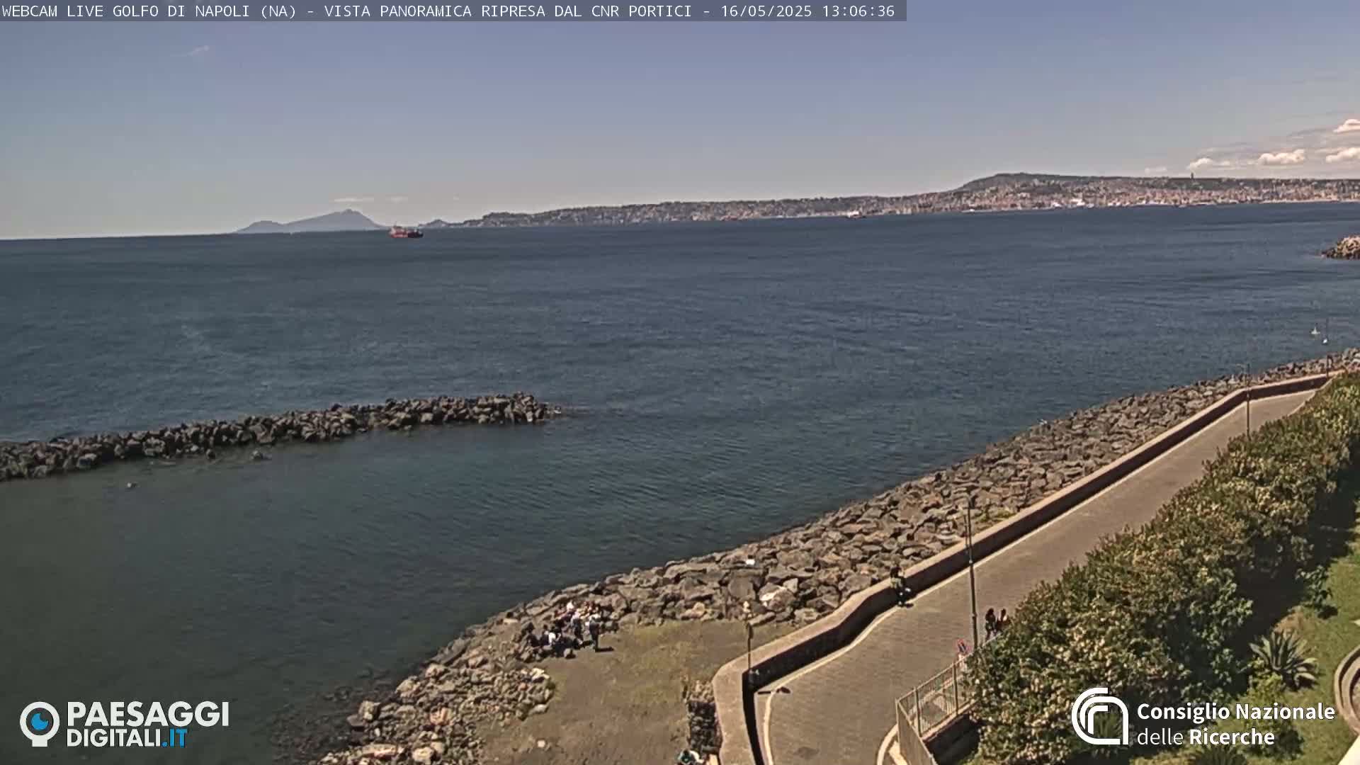 A rocky shoreline curves gently alongside a paved walkway, overlooking a calm bay with a distant mountain range under a clear, sunny sky.