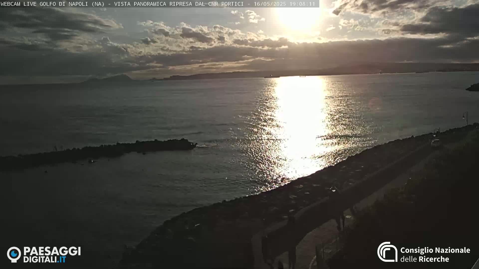 A partly cloudy sunset casts shimmering light on a calm sea, with a rocky shoreline and distant land visible.