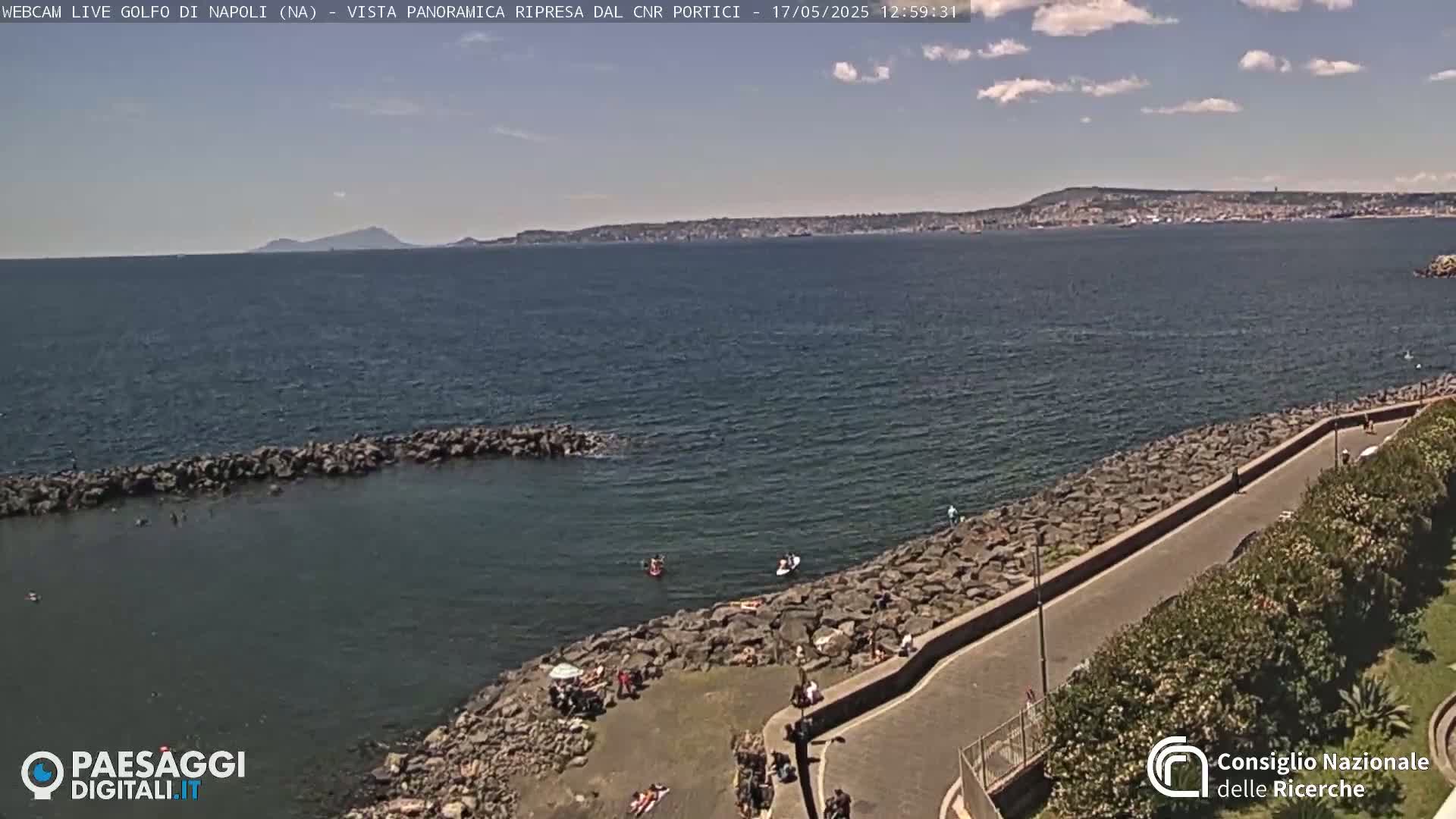 A partly cloudy day shows a rocky shoreline with people sunbathing and swimming in calm water, overlooking a city and a distant mountain range.