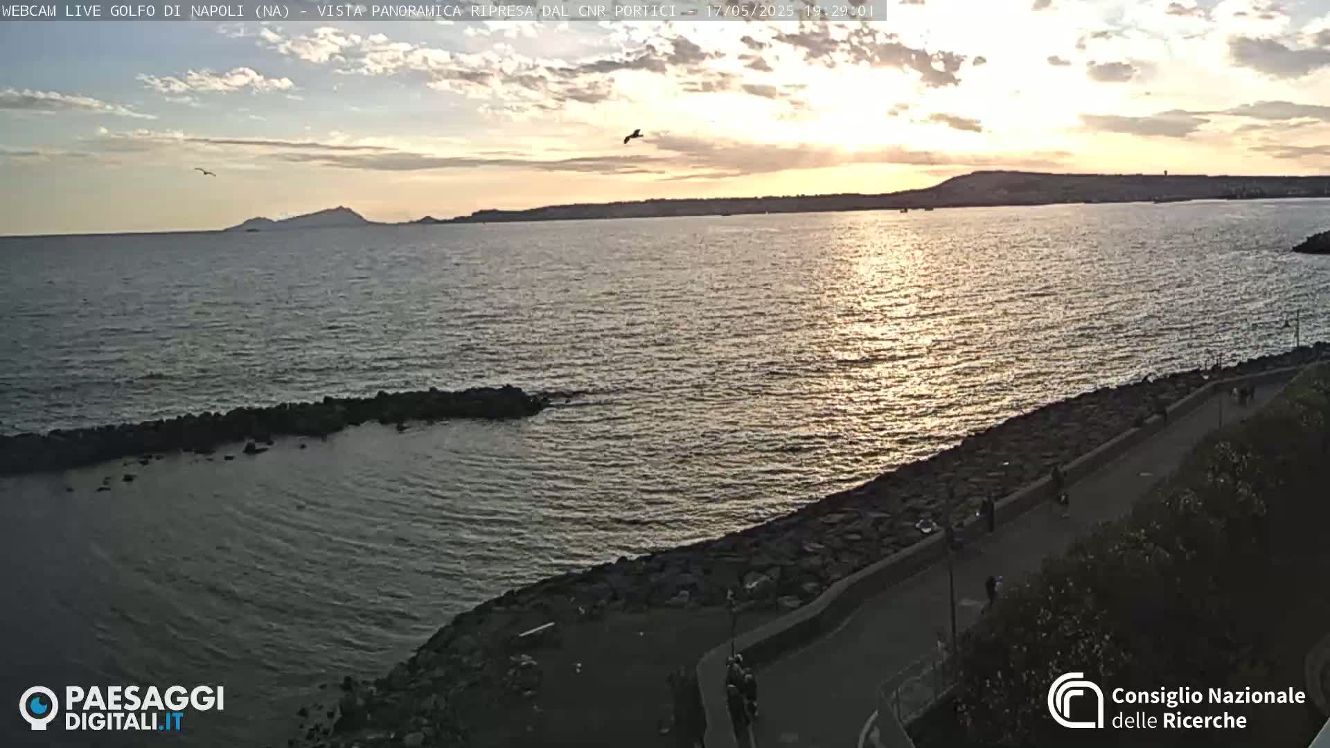 A calm sea at sunset is viewed from an elevated position, showing a rocky shoreline with a path and a few people walking along it, and a distant landmass under a partly cloudy sky.