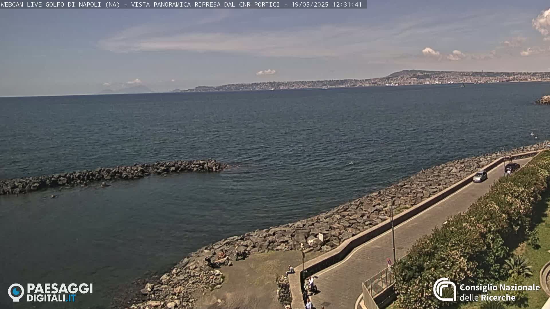 A partly cloudy day shows a rocky shoreline with a road curving along the water's edge, overlooking a calm sea and a distant city skyline under a mostly clear sky.