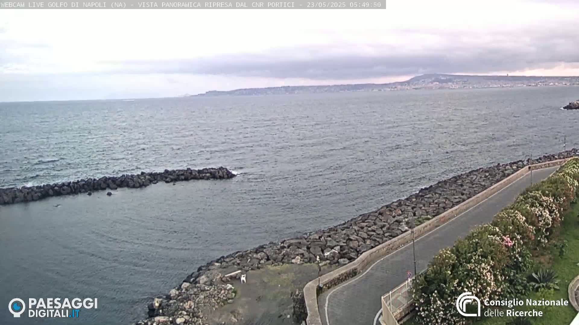 A rocky shoreline curves gently around a calm sea under a cloudy sky, with a distant cityscape and mountain visible on the horizon.