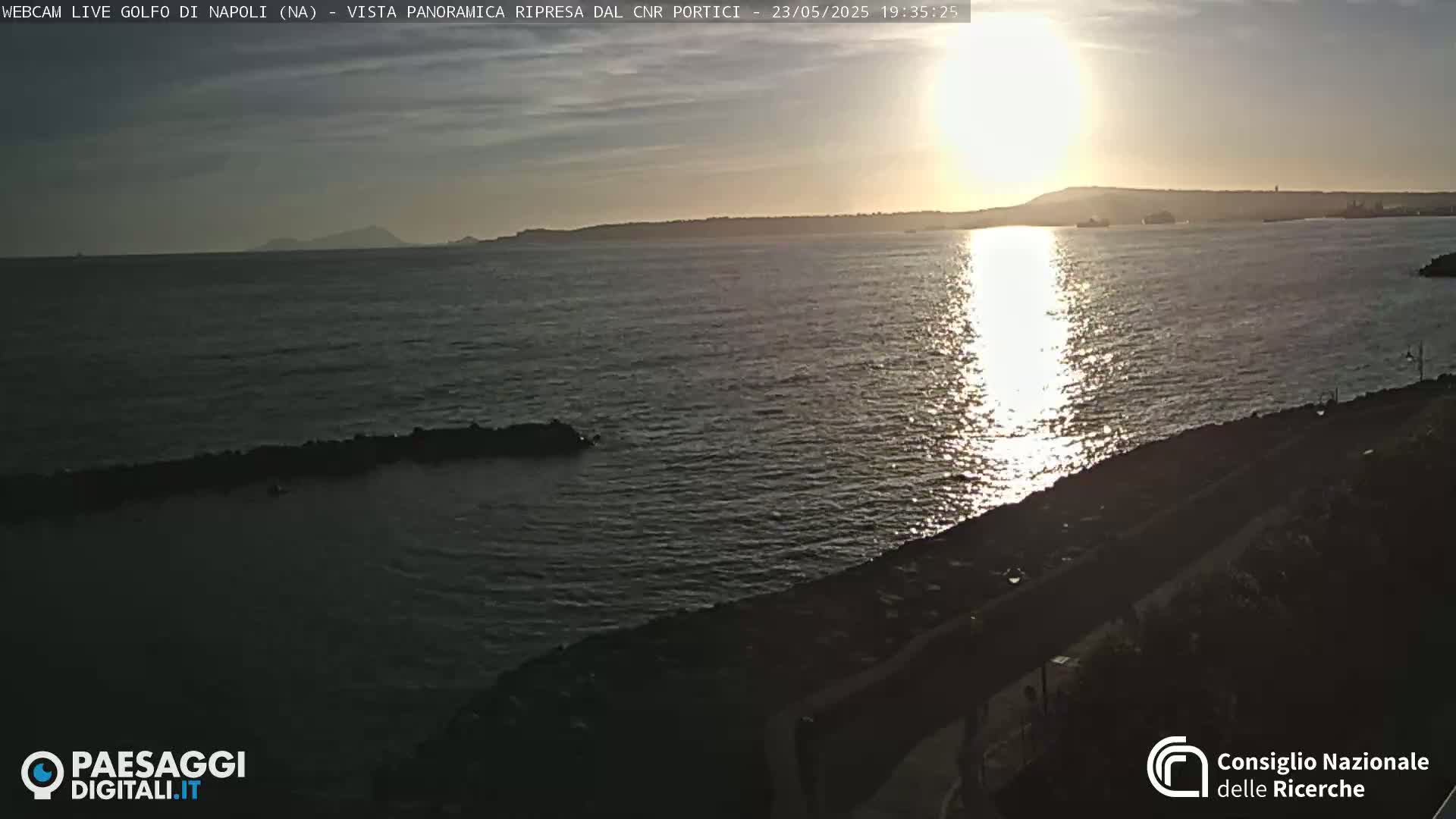A calm sea reflecting the setting sun, with a distant landmass visible on the horizon under a mostly clear sky.
