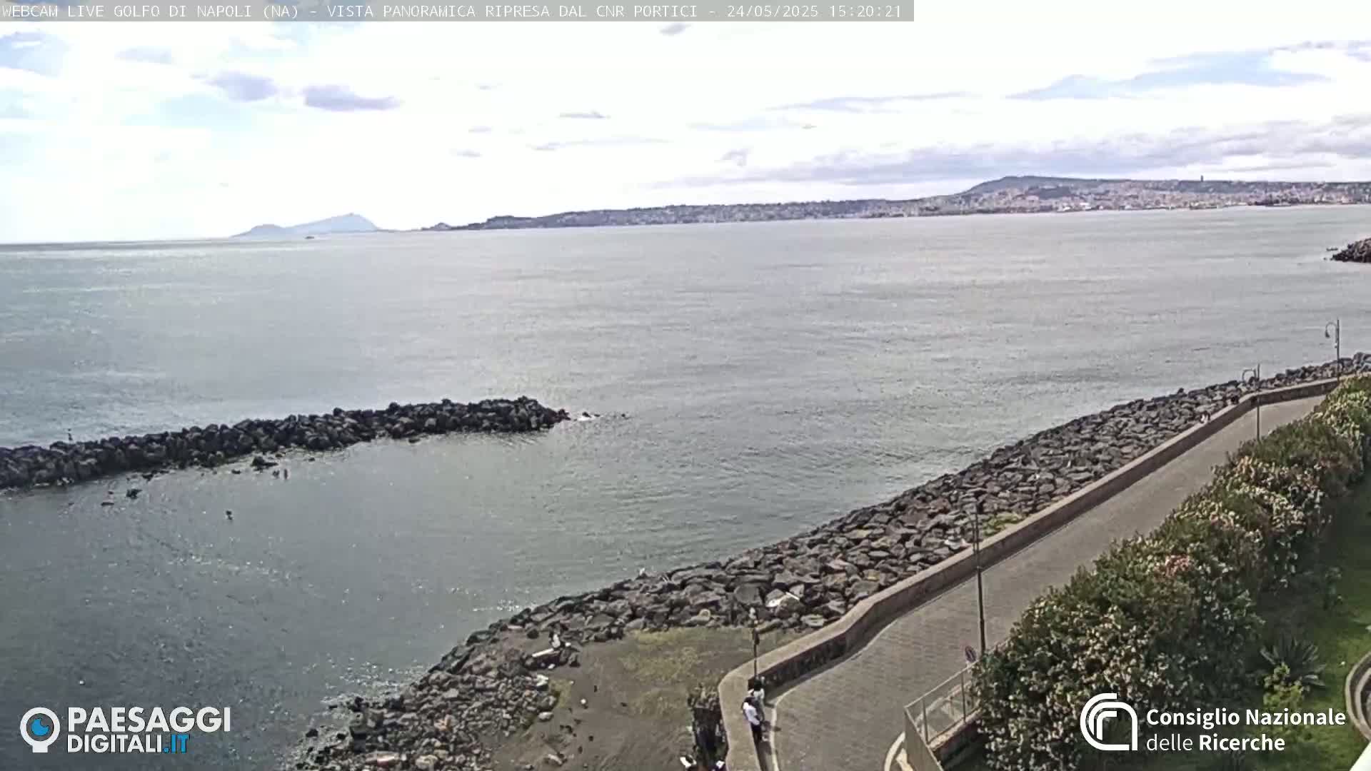 A rocky shoreline with a paved walkway curves alongside calm, gray-blue water under a partly cloudy sky, with a distant city and mountain visible on the horizon.