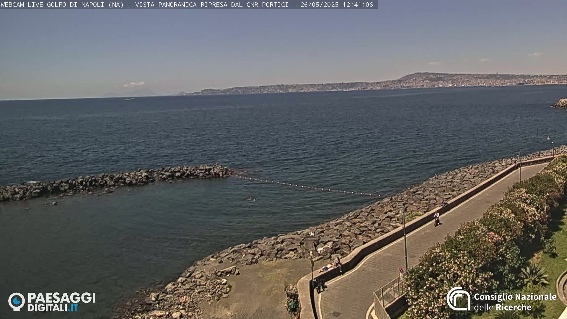 A rocky shoreline with a paved path and vegetation overlooks a calm sea under a clear sky, with a distant cityscape and mountains visible on the horizon.