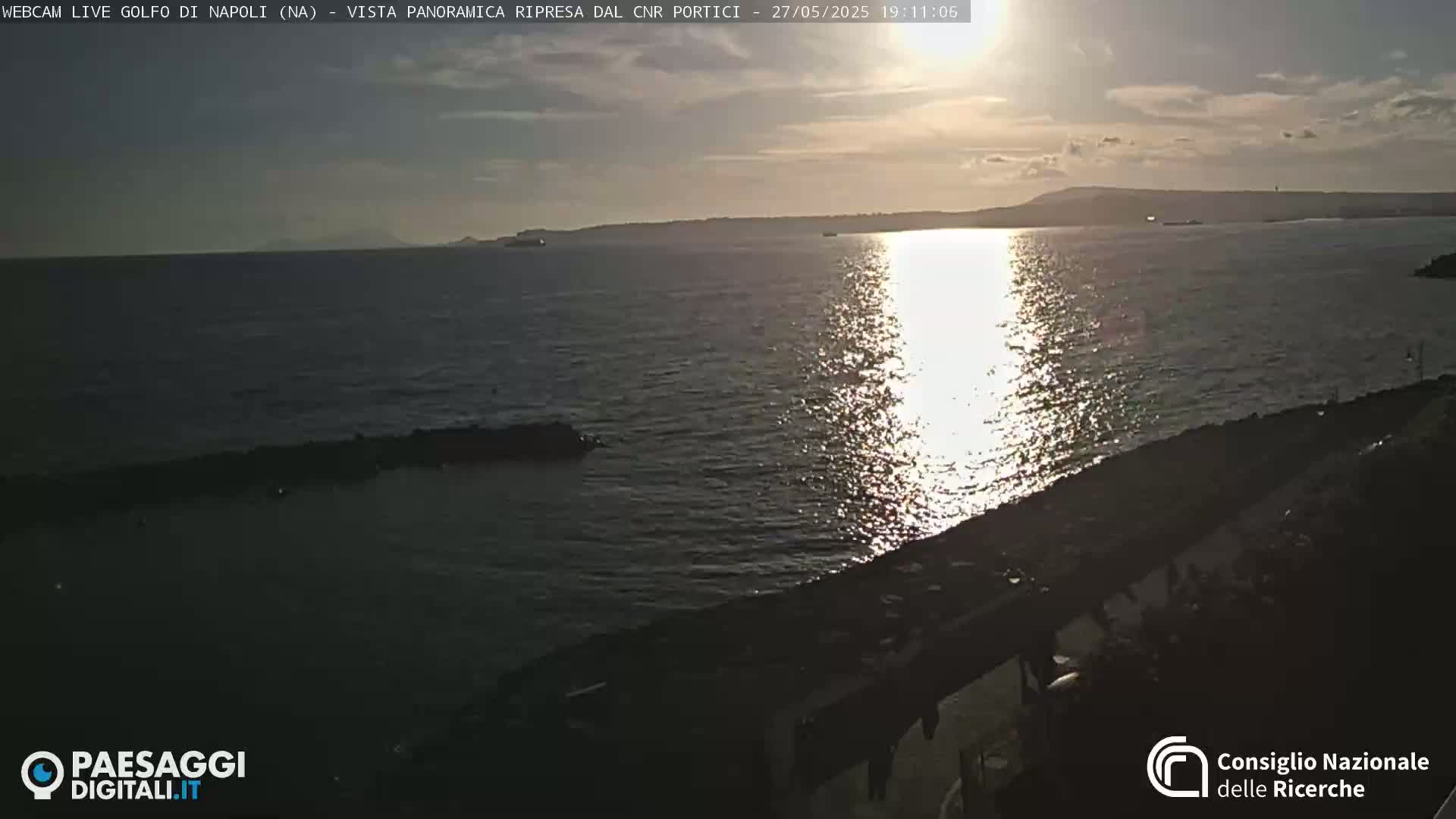 A calm sea reflects the setting sun, with a distant shoreline visible under a partly cloudy sky.