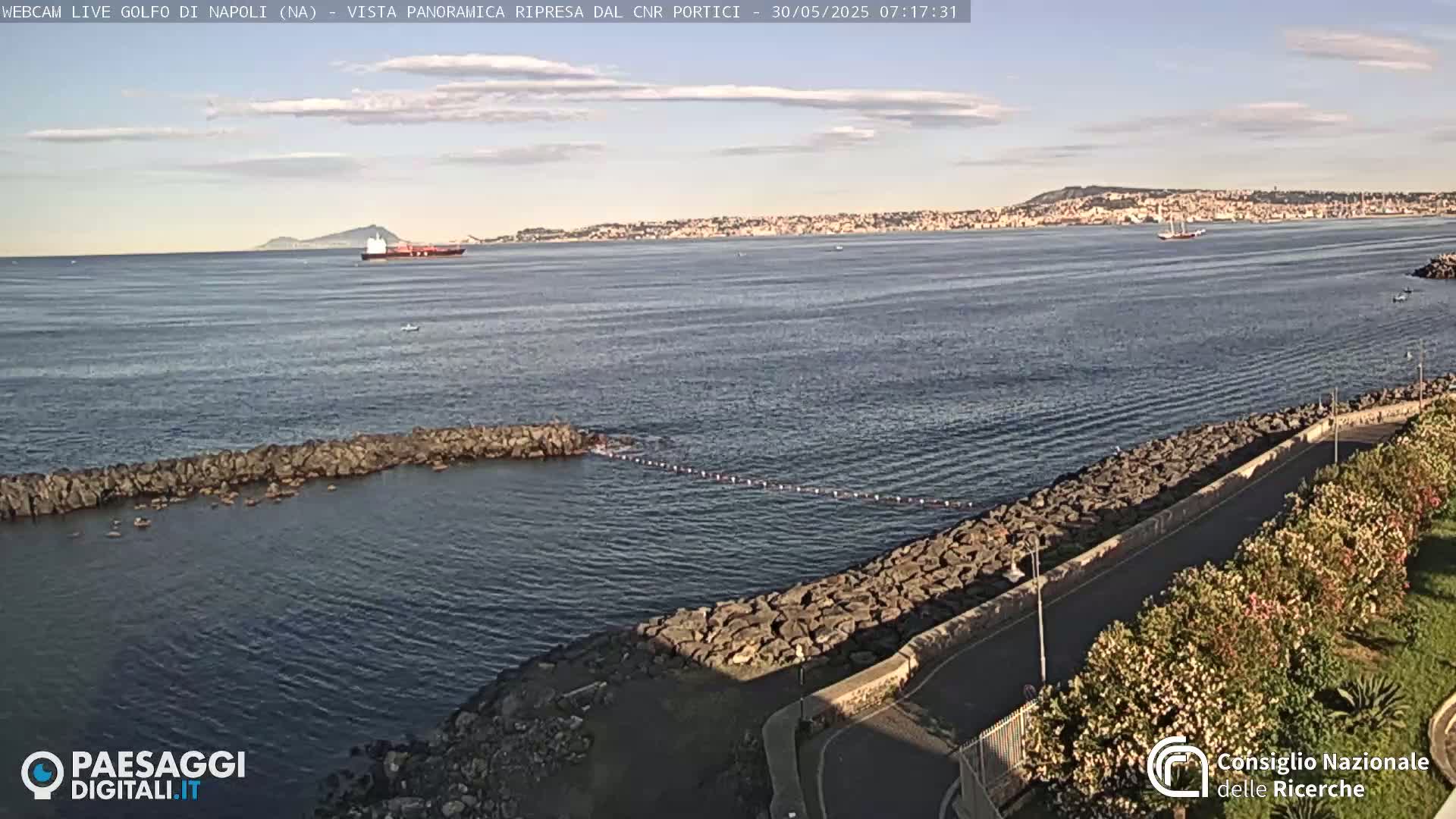 A partly cloudy day shows a calm bay with a rocky shoreline, a distant cityscape, and several ships on the water.