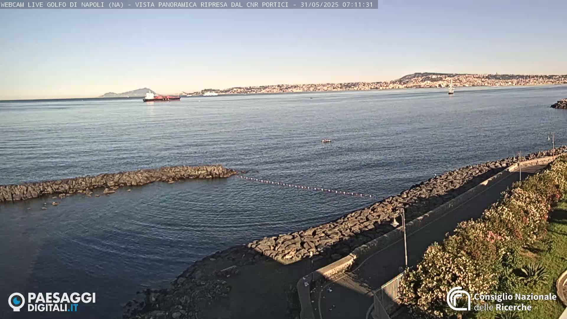 A calm sea with a rocky shoreline and distant cityscape under a clear sky is visible from a high vantage point.