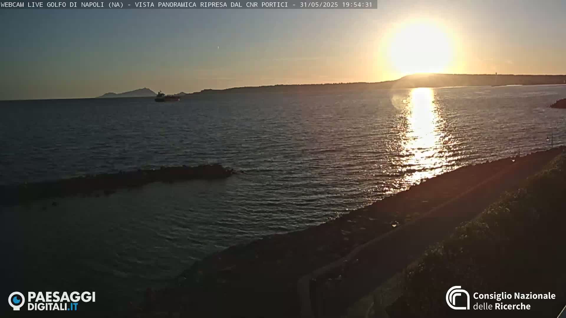 A calm body of water reflects the setting sun, with a distant island and a small vessel visible on the horizon.
