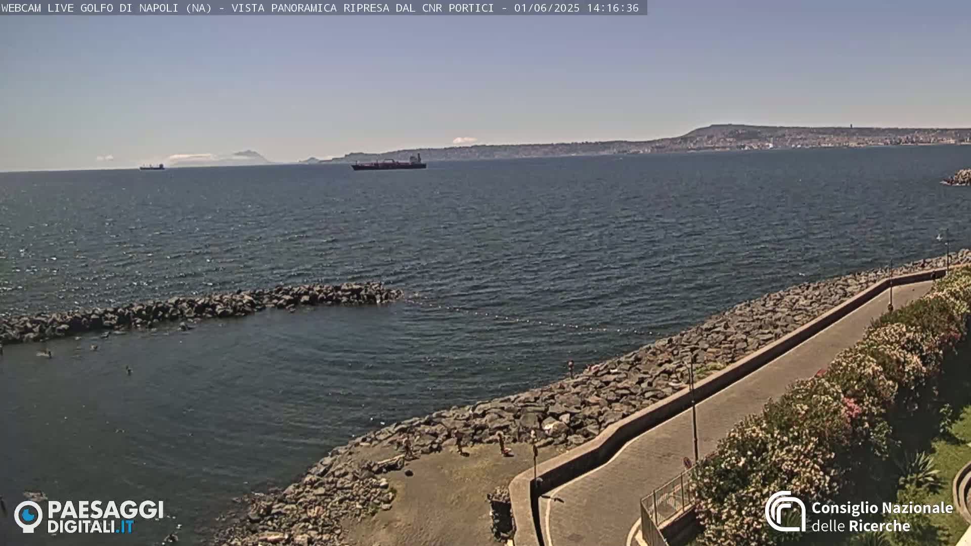 A rocky shoreline with a paved walkway curves into the frame, overlooking a calm sea where a large ship is visible on the horizon under a clear sky.