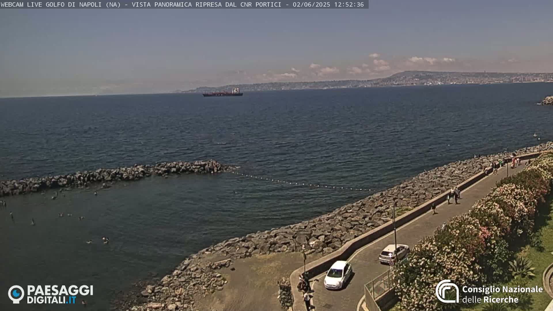 A rocky shoreline with people walking along a path overlooking a calm sea, a distant ship, and a partly cloudy sky.