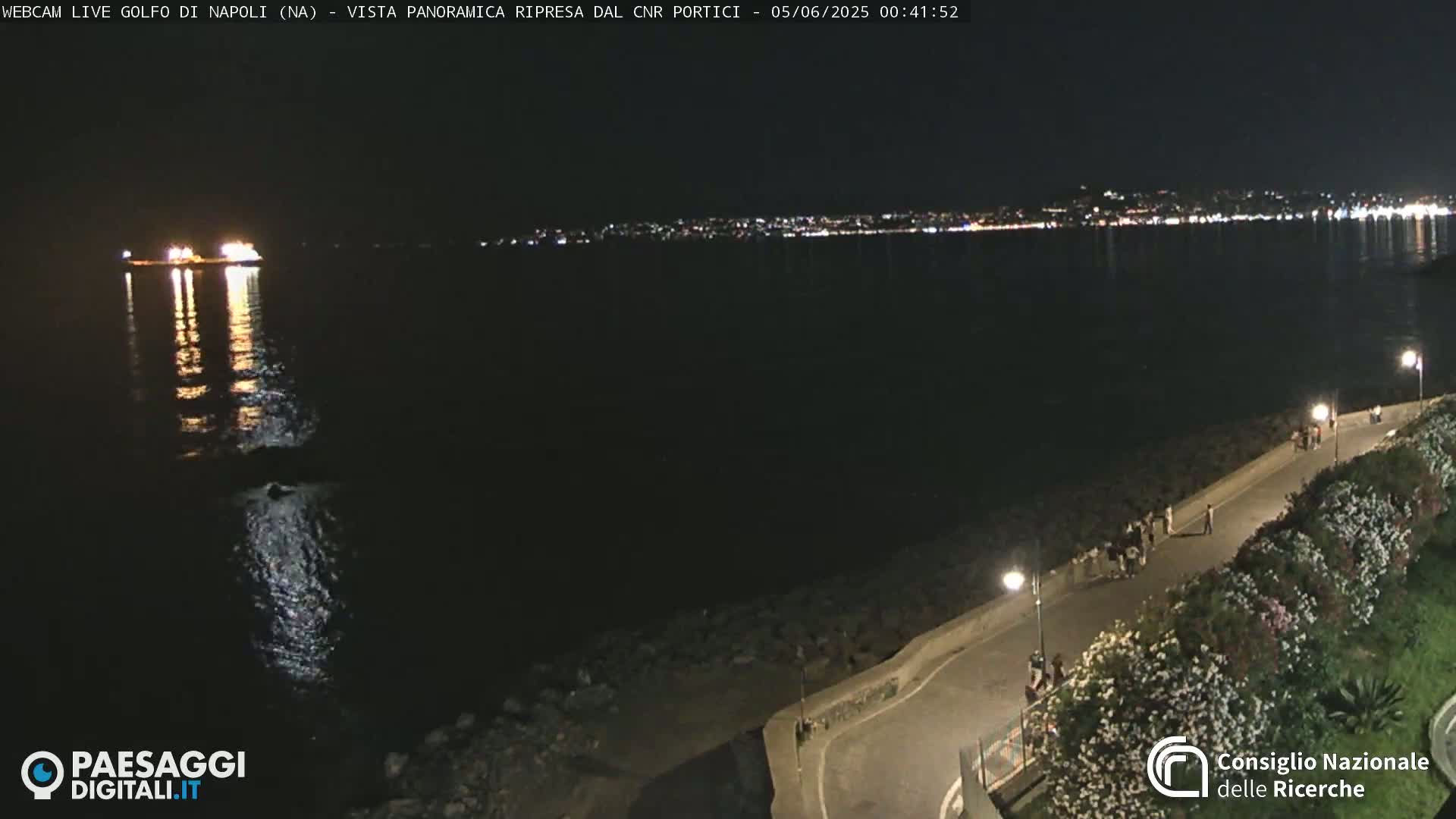 A nighttime view of a coastline, featuring a dark body of water reflecting city lights in the distance, with a few people walking along a lighted path beside the water.