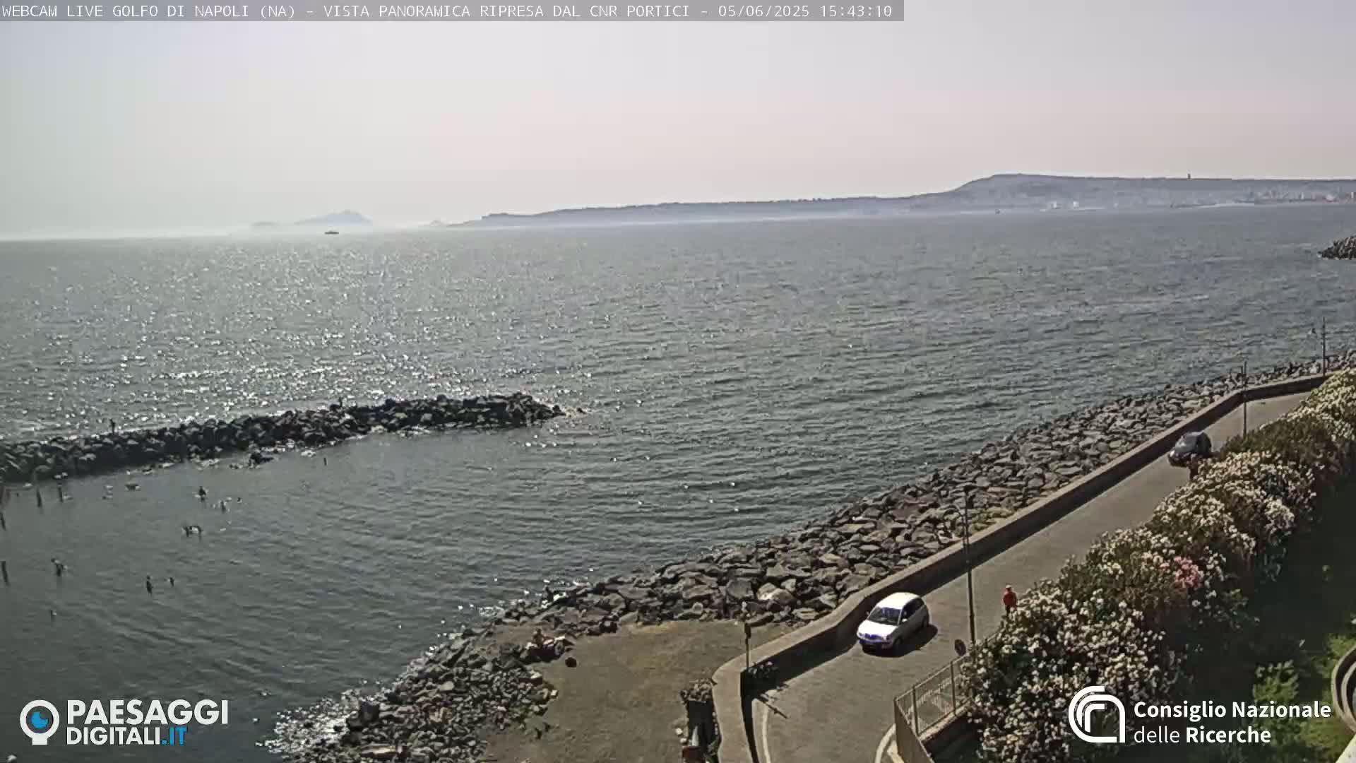 A rocky shoreline with a road and vegetation overlooks a calm sea under a clear, sunny sky; a distant landmass is visible on the horizon.