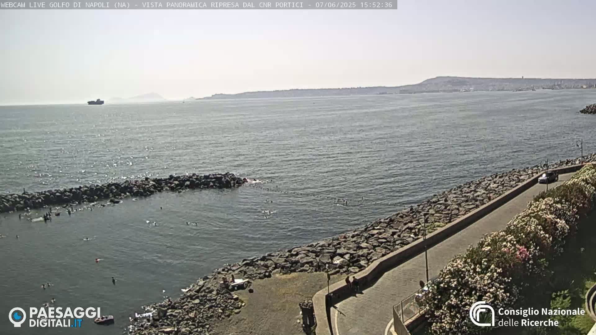 A rocky shoreline with people swimming in the calm sea under a sunny sky, a distant coastline visible in the background.