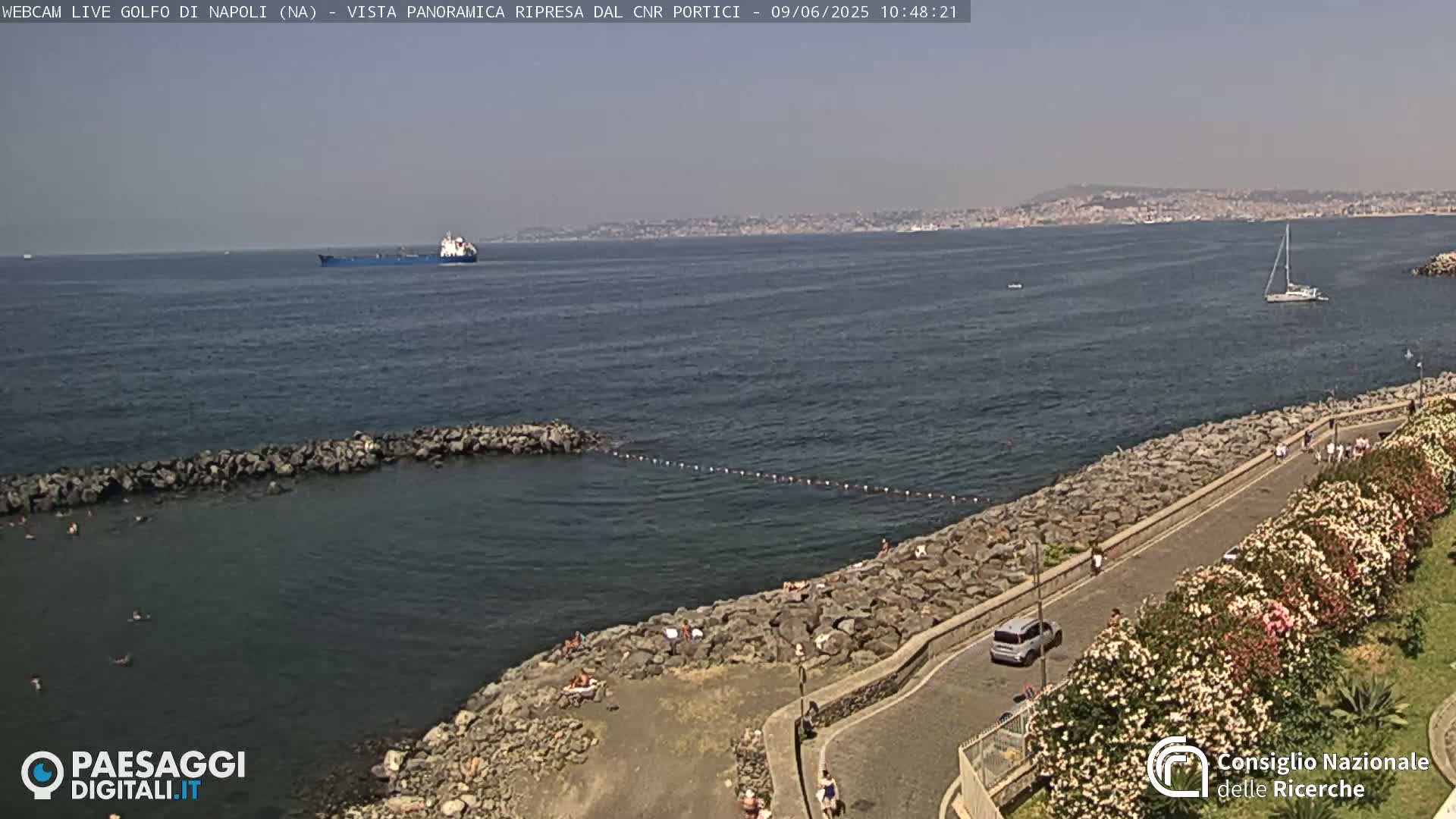 A rocky shoreline with people sunbathing and a road lined with flowering bushes overlooks a calm sea with a large cargo ship and a sailboat in the distance under a hazy, sunny sky.