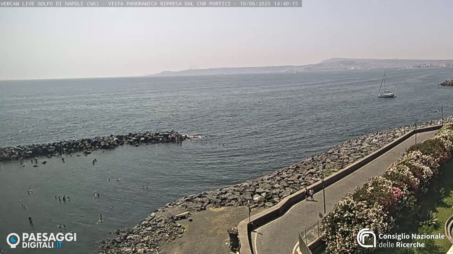 A rocky shoreline with people swimming in calm, clear water, a paved walkway lined with flowering bushes, and a sailboat visible in the distance under a bright, sunny sky.