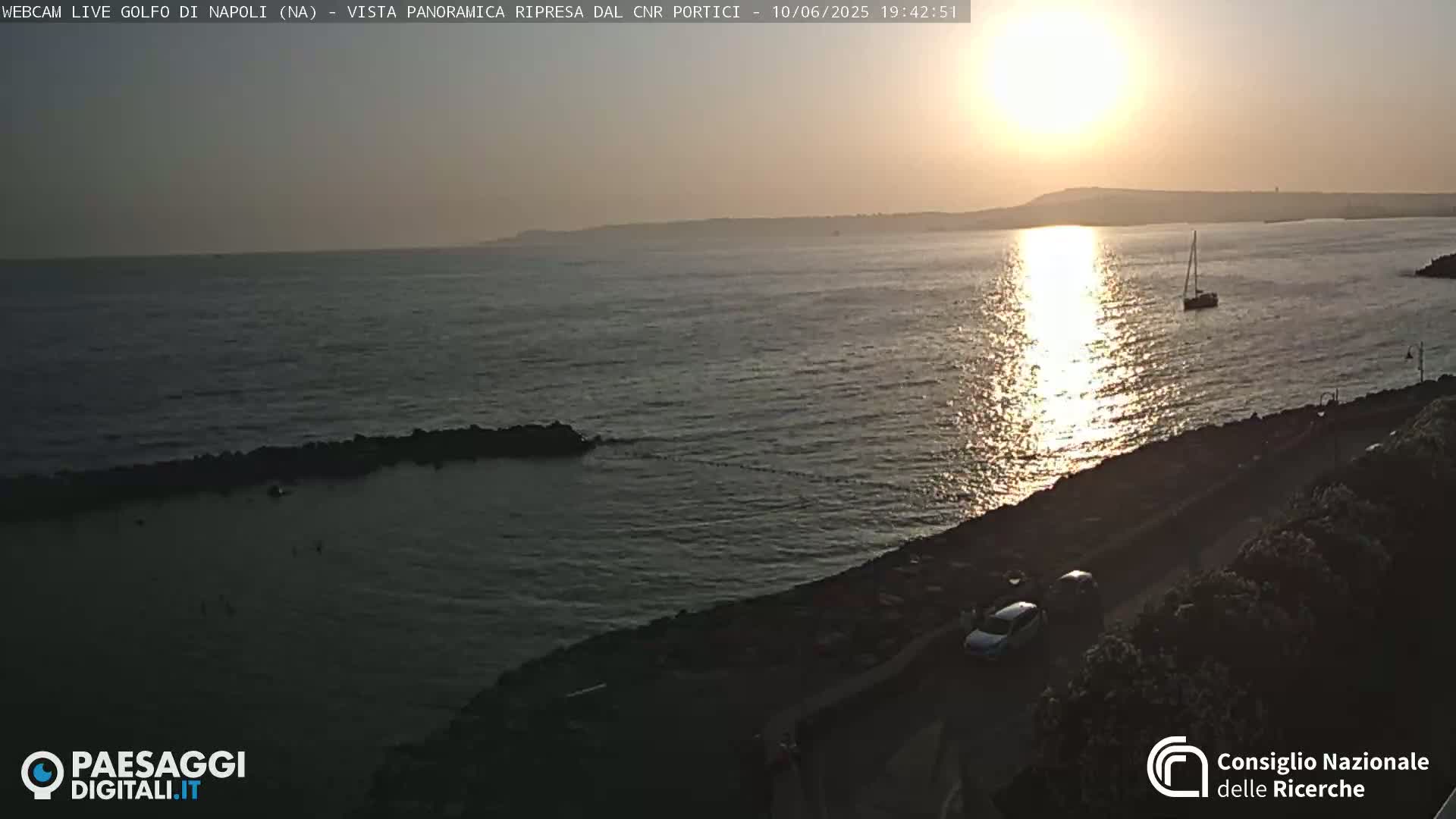 A sunset over a calm body of water, with a sailboat visible in the distance, and a few cars parked along a rocky shoreline.