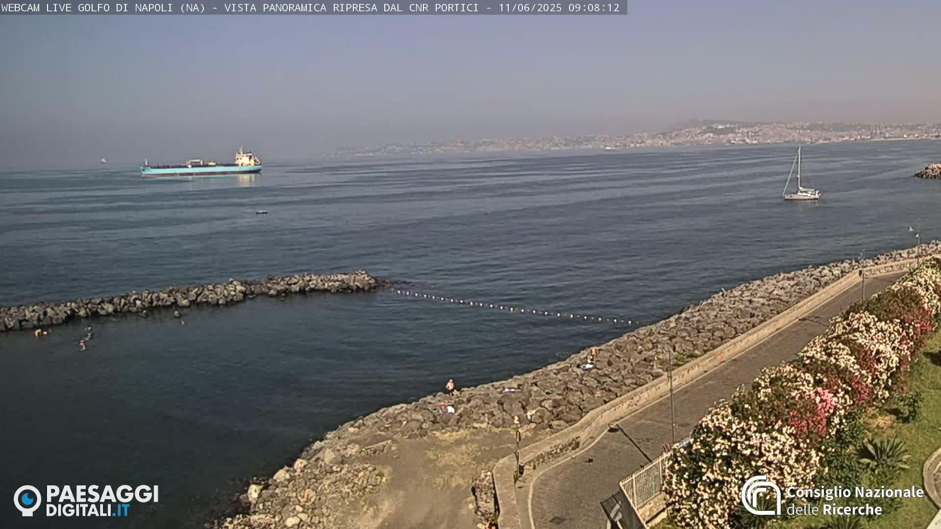 A sunny day shows a rocky shoreline with people swimming in calm water; a cargo ship and sailboat are visible in the bay, with a distant cityscape in the background.