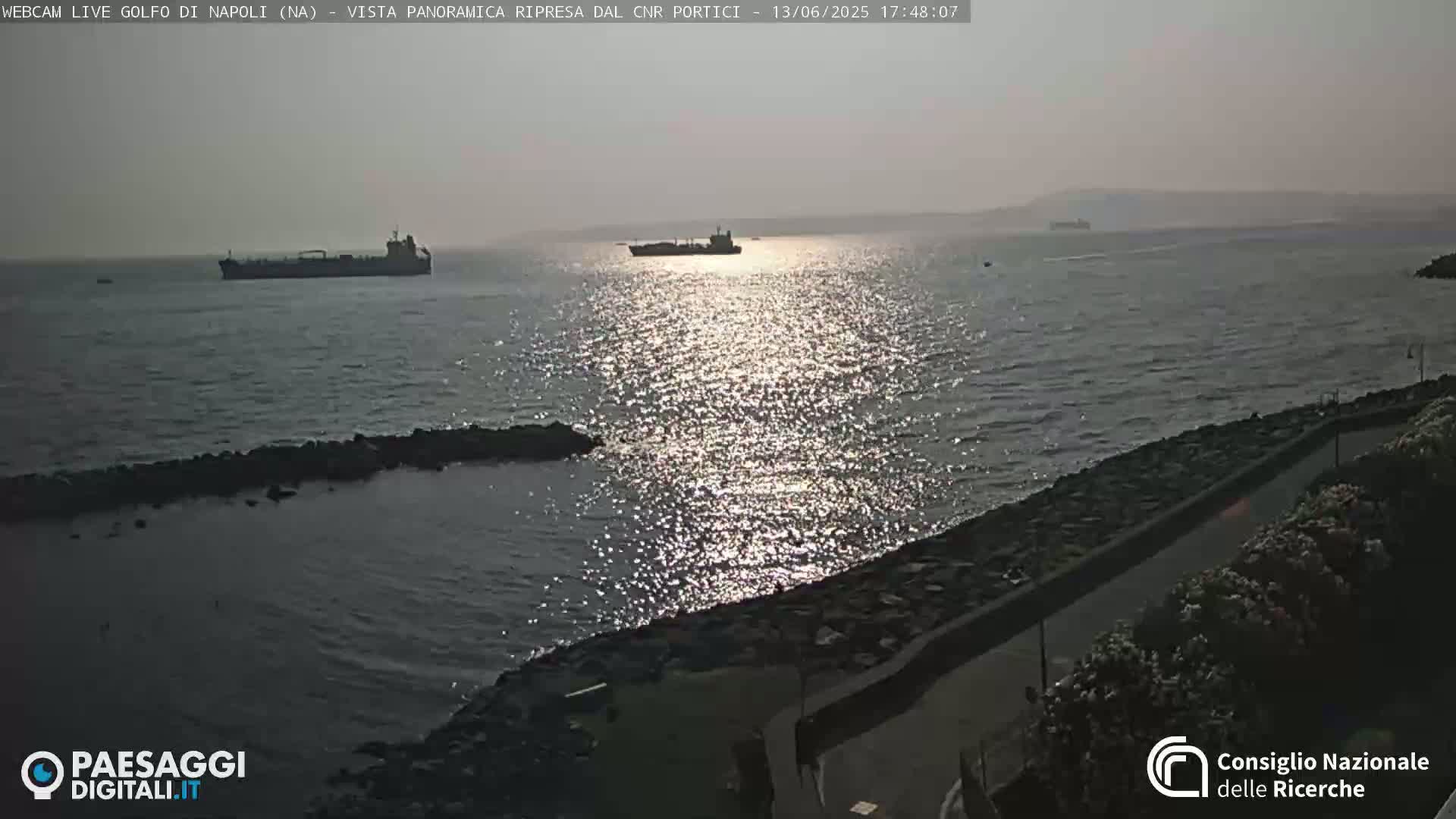 A calm, sunlit sea with several cargo ships in the distance is viewed from a rocky shoreline with a road visible on the right.