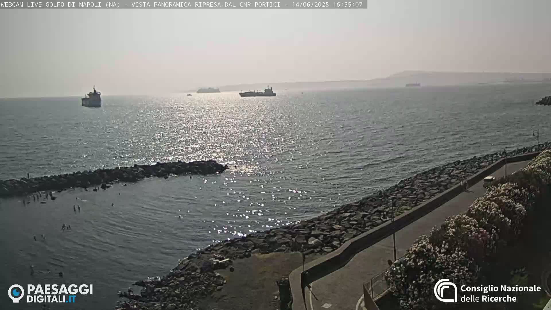 A hazy, sunny day shows a calm sea with several ships in the distance, a rocky shoreline with people swimming near a small, protected cove, and a paved road lined with vegetation.
