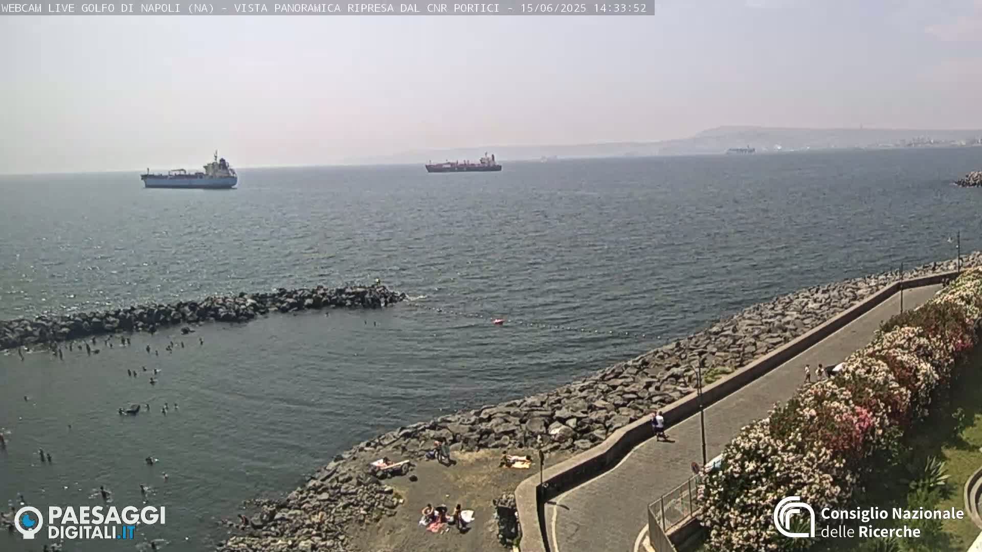A hazy, sunny day shows a rocky shoreline with people sunbathing and walking along a paved path, while several ships are visible on the calm sea in the distance.
