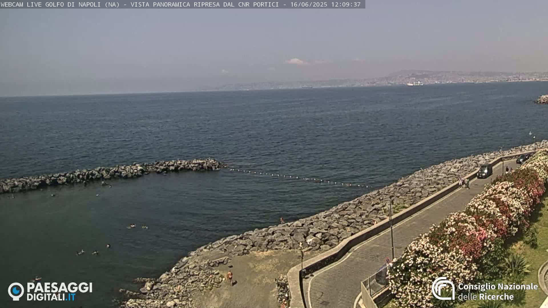 A rocky coastline with a calm sea stretches to a hazy distant shoreline under a partly cloudy sky.