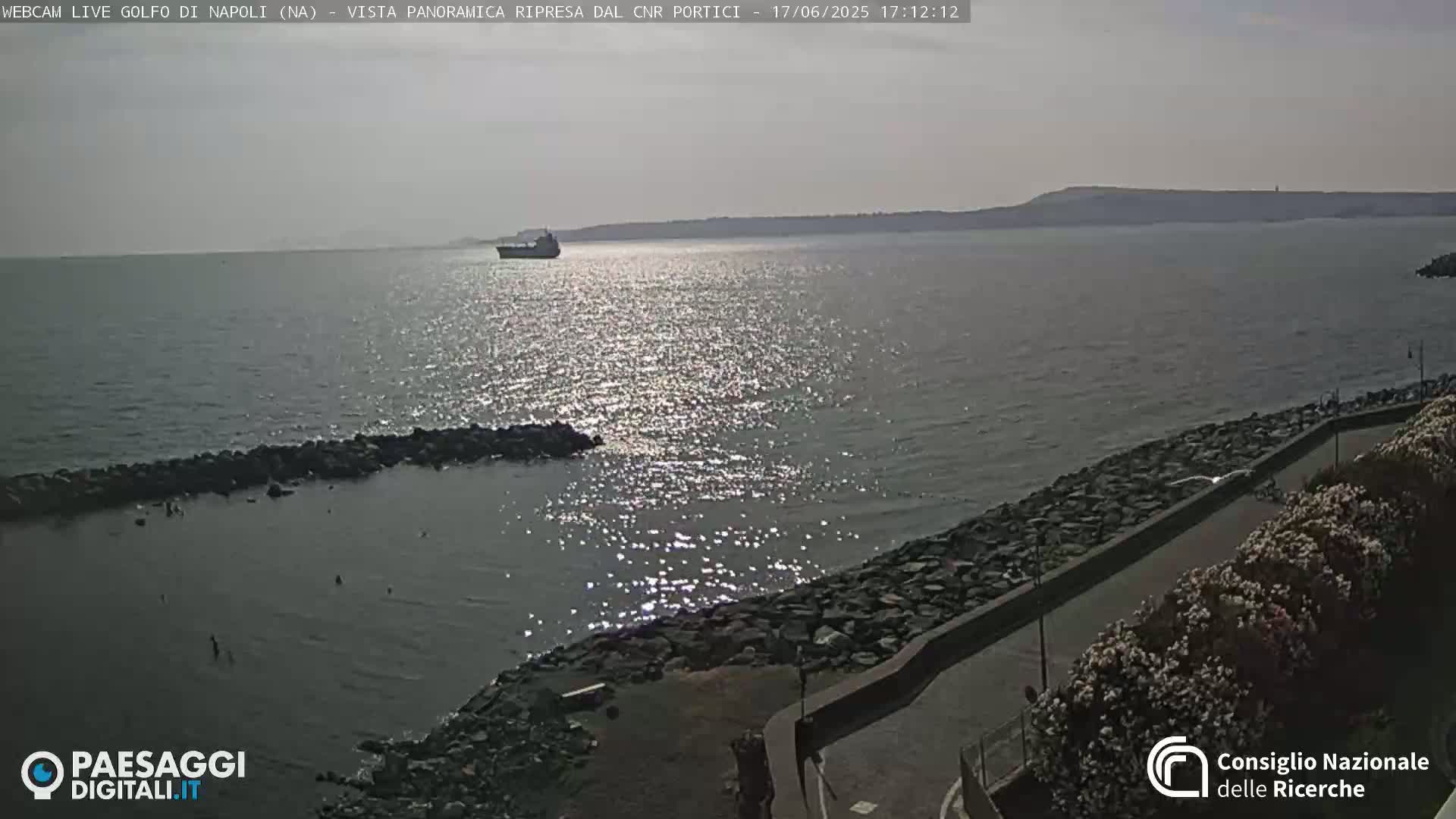 A calm body of water with a distant ship and a rocky shoreline under a bright, hazy sky.