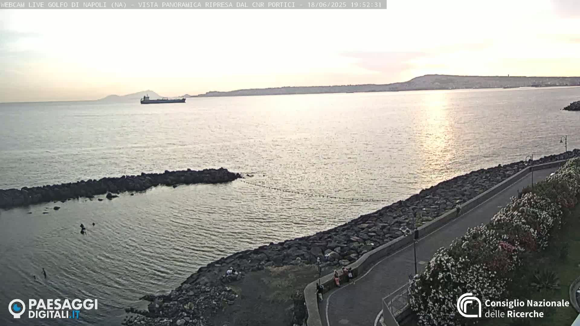 A calm sea with a rocky shoreline and a distant cargo ship under a twilight sky.
