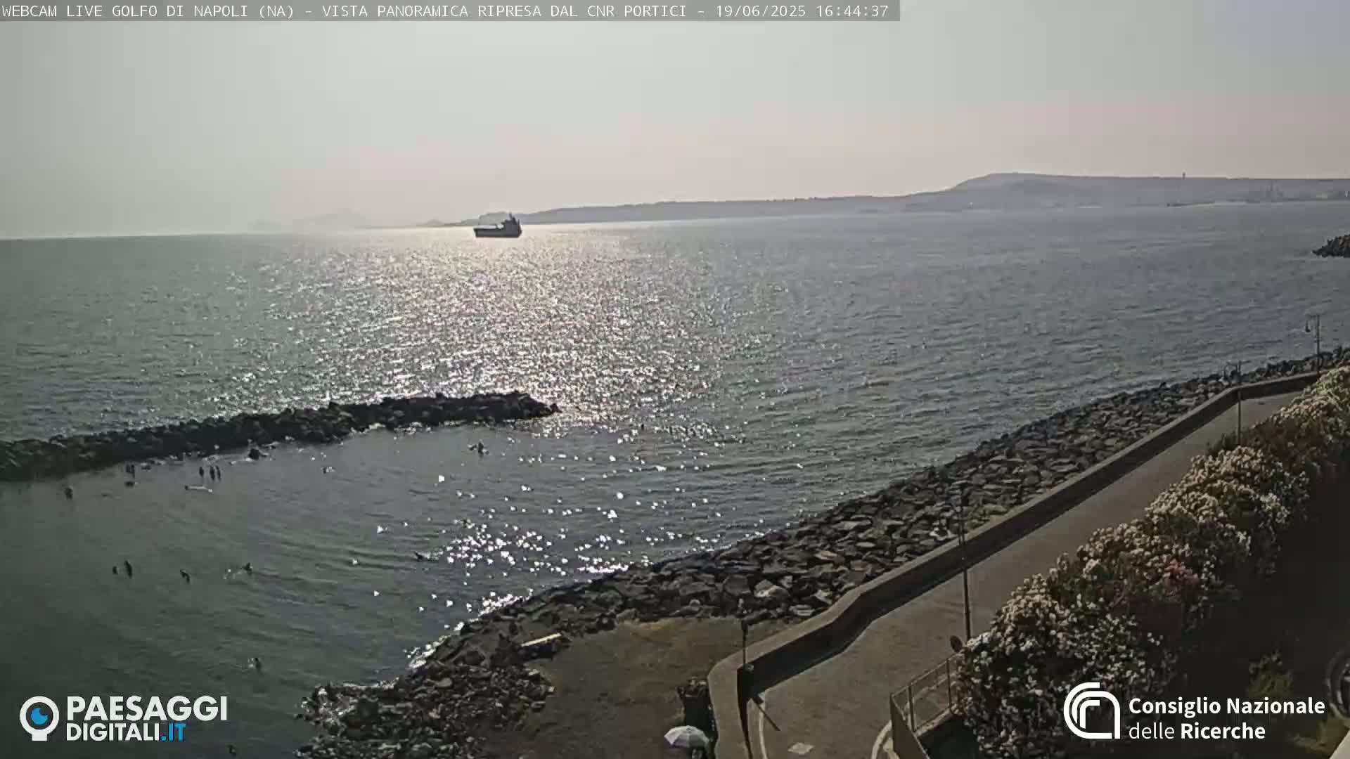 A sunny day shows a calm sea with a few people swimming near a rocky shoreline and a distant ship.