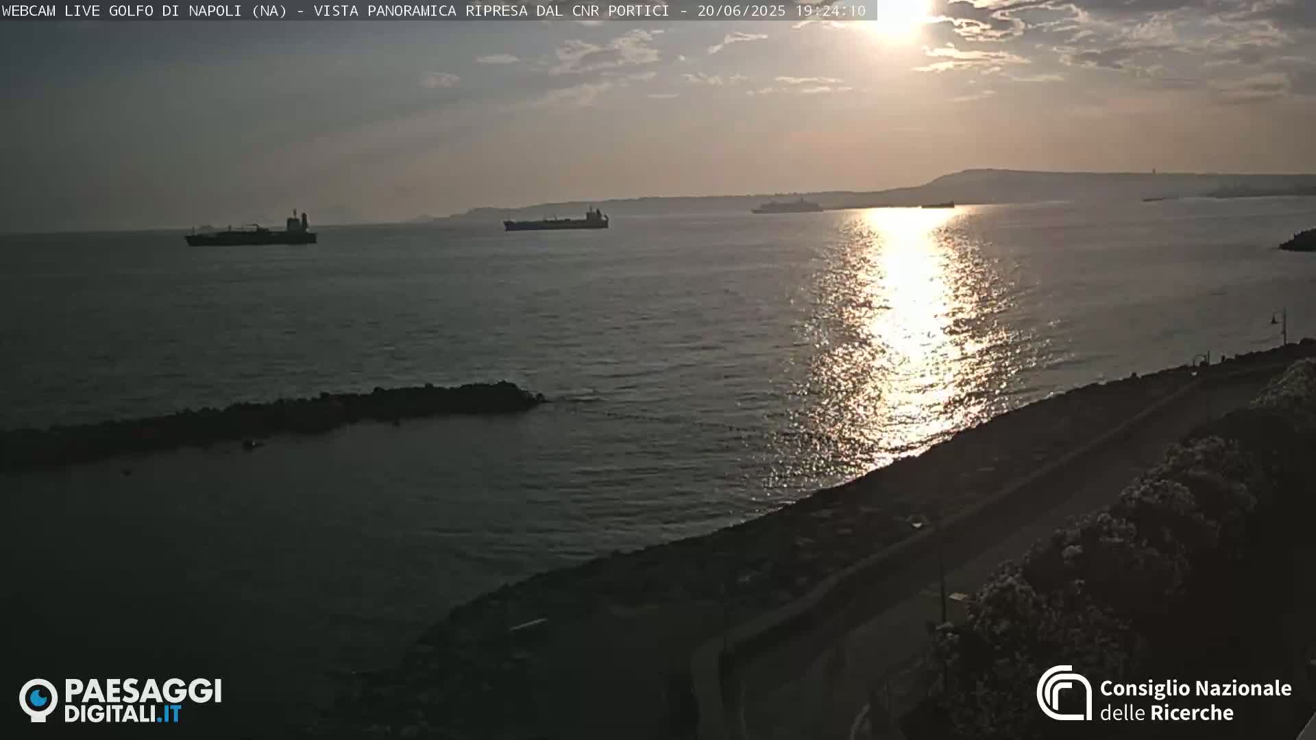 Several ships are on a calm sea at sunset, viewed from a shoreline with a roadway.