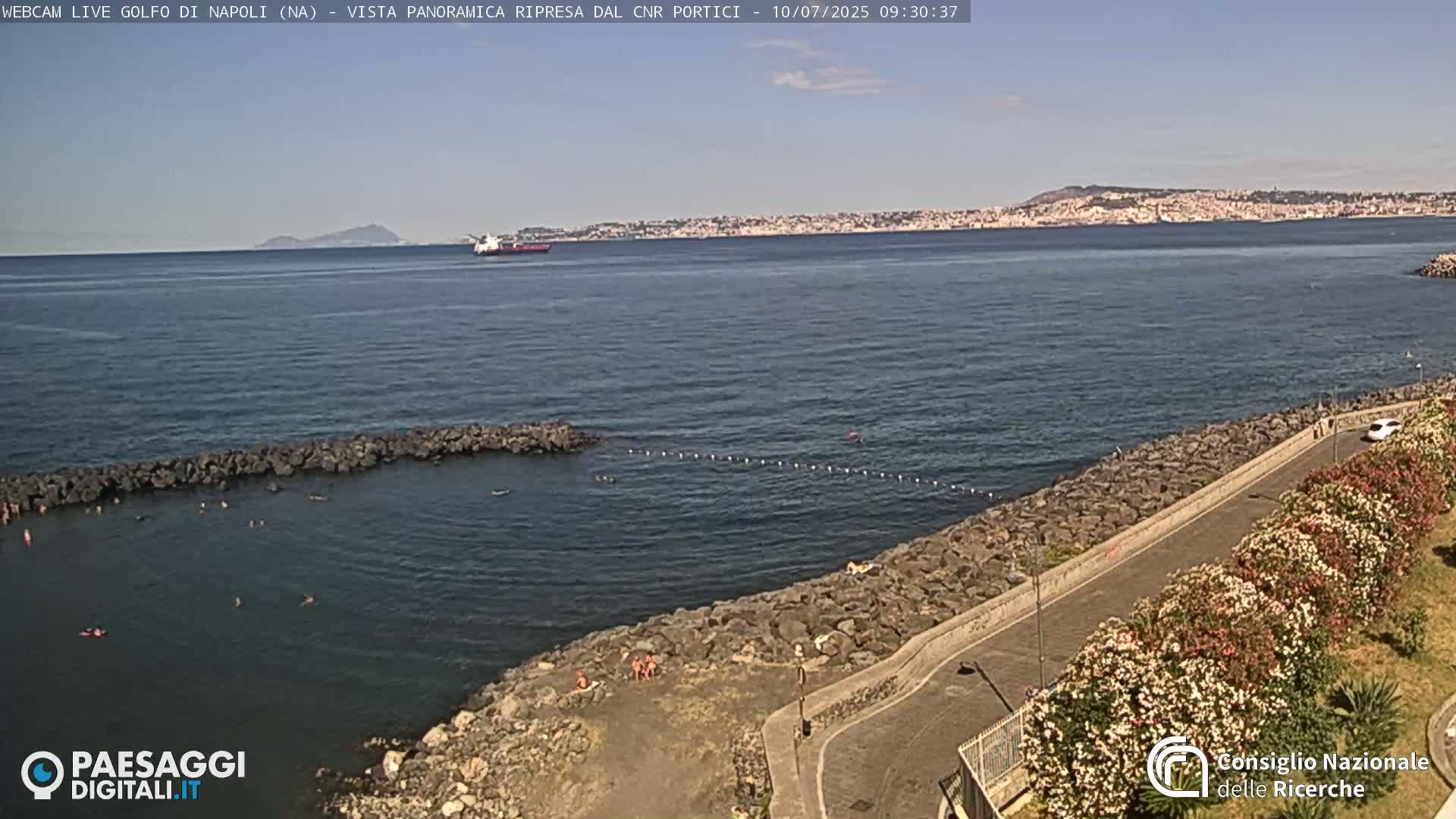 A rocky shoreline with people swimming in a calm, clear ocean, a city skyline and distant mountains visible in the background under a sunny sky.