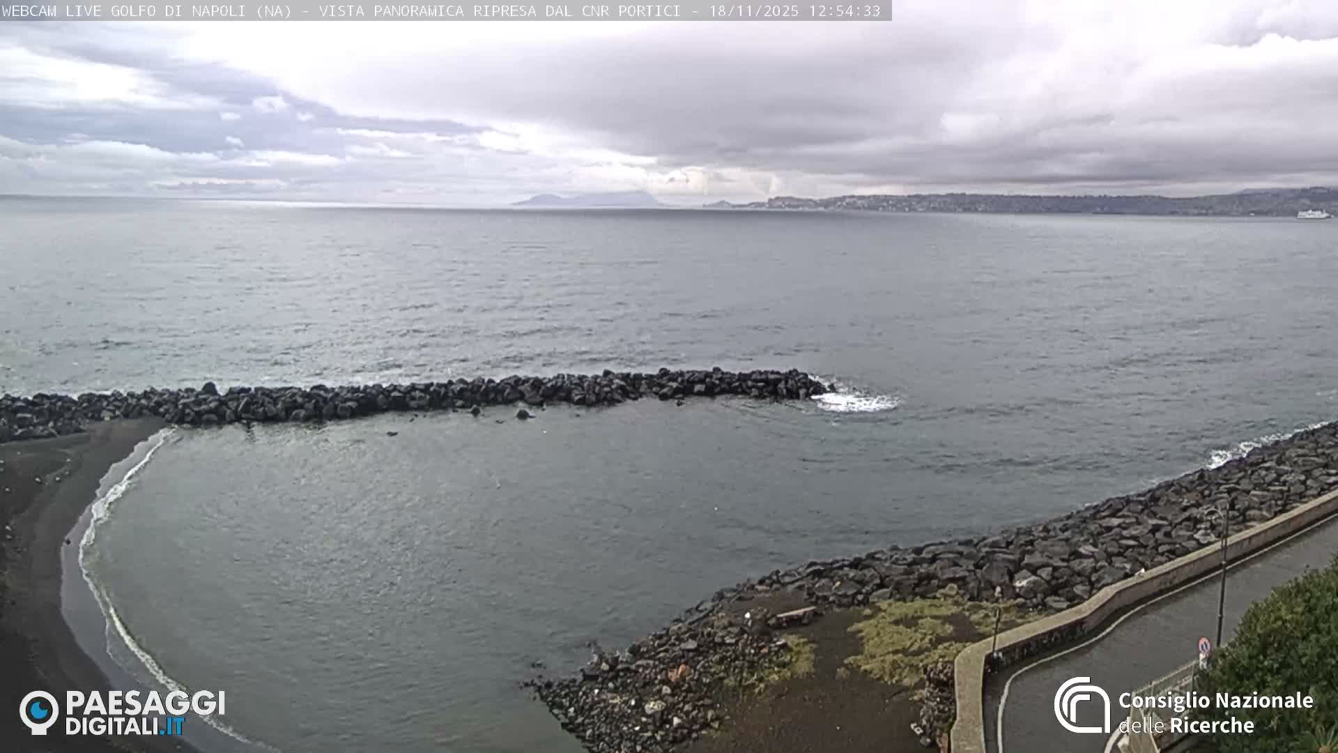 A dark, clear night scene reveals a curved coastal road illuminated by a single streetlamp, overlooking calm water with the faint glow of a distant city on the horizon.