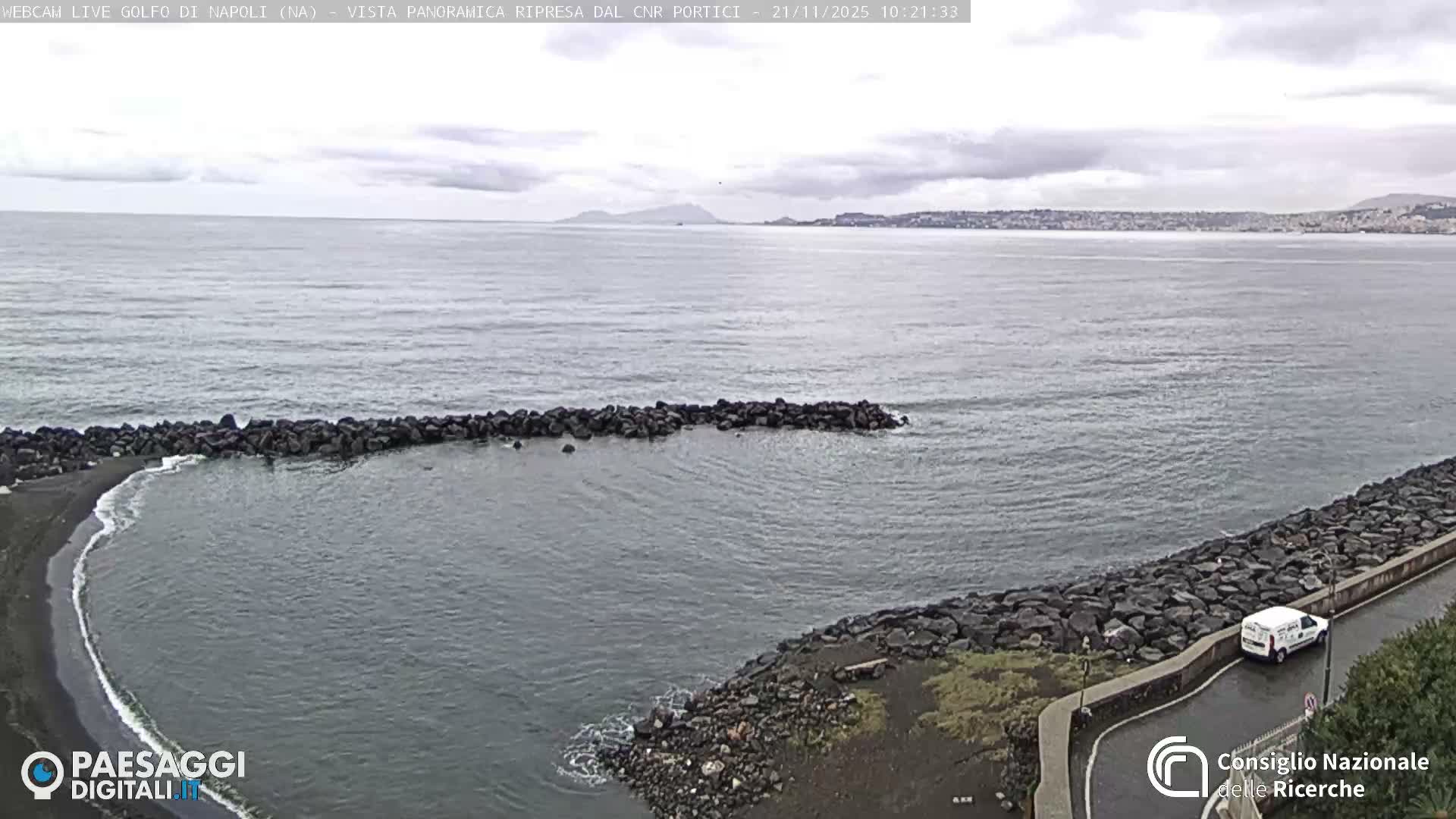 A wide view shows a calm, gray sea under an overcast sky, with a dark rocky beach and breakwater in the foreground, and a wet coastal road with a white van parked on it.