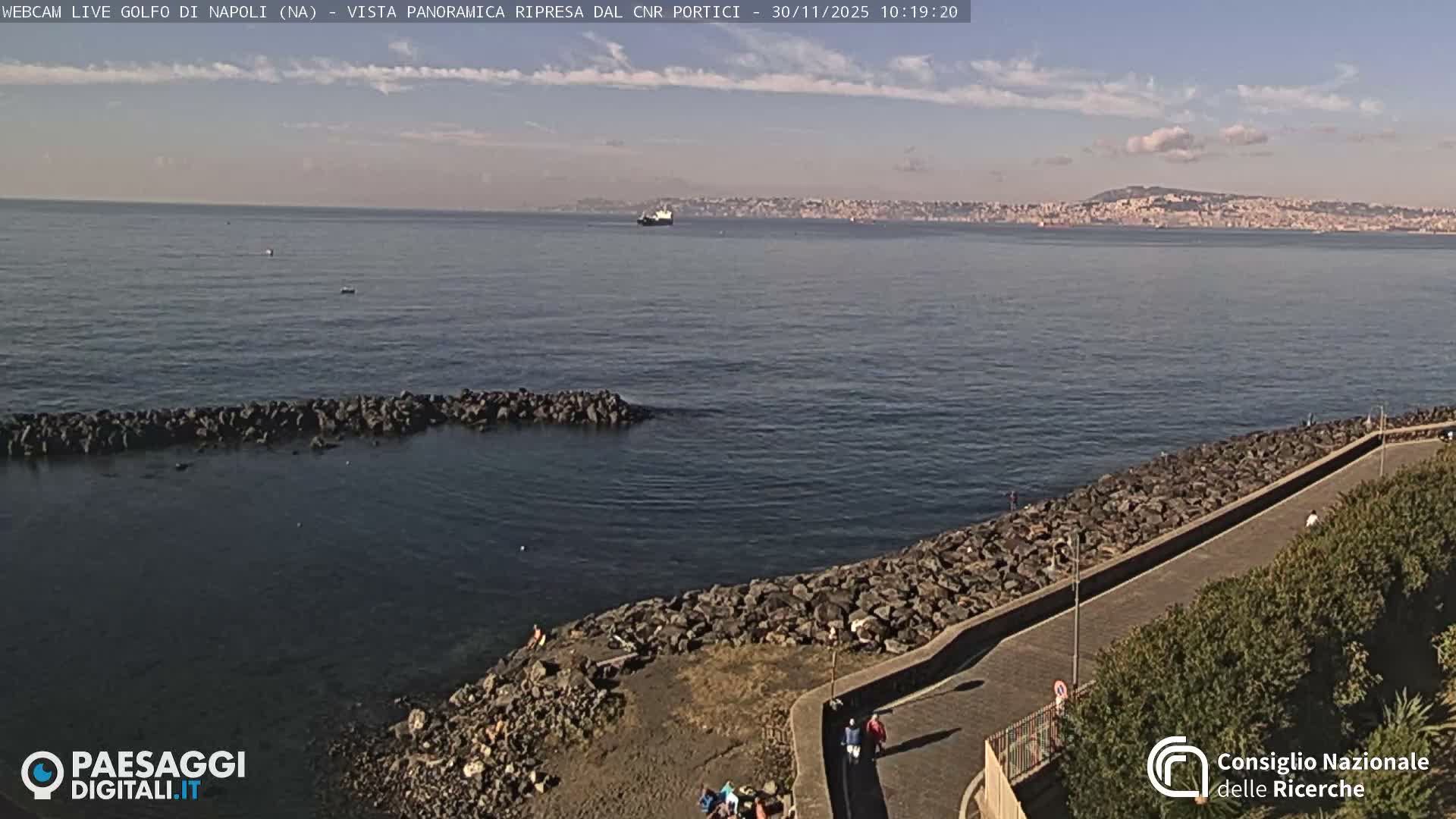 A panoramic view captures a calm sea with a rocky breakwater and a paved path in the foreground where people stroll, while boats navigate the water towards a distant city nestled against hazy hills, all under a bright, partly cloudy sky.