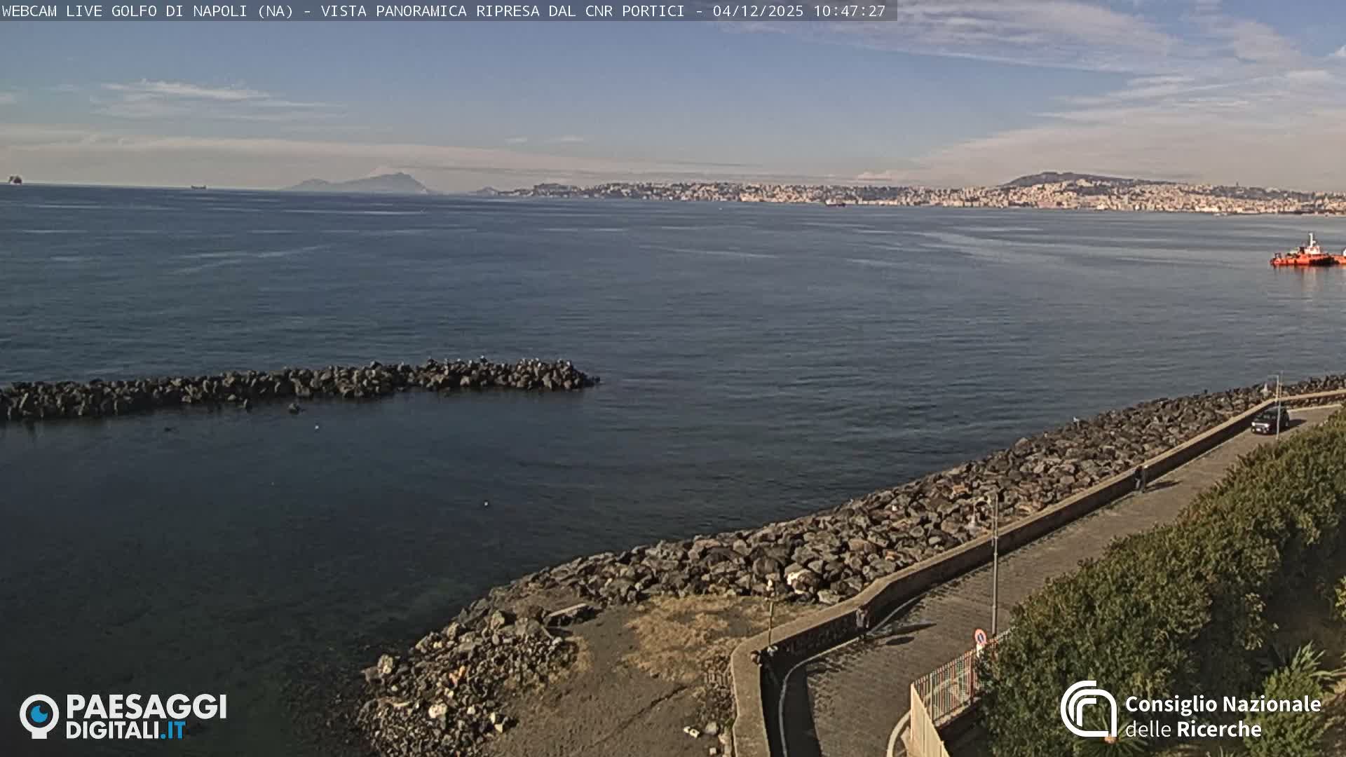 A panoramic view captures a calm bay with a rocky shoreline and a paved promenade on the right, leading to a distant city and mountains under a bright, partly cloudy sky, with a few boats visible on the water.