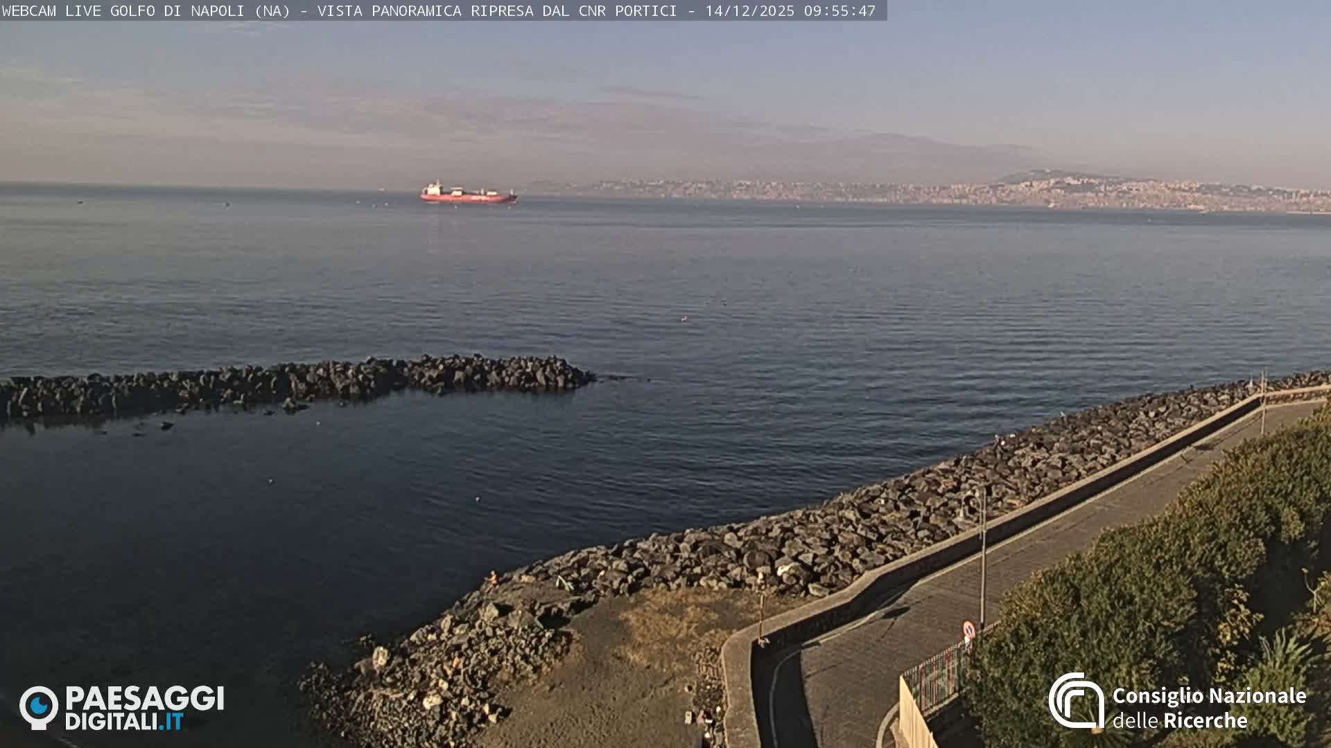 A panoramic view captures a calm bay with a rocky shoreline and a paved promenade on the right, leading to a distant city and mountains under a bright, partly cloudy sky, with a few boats visible on the water.