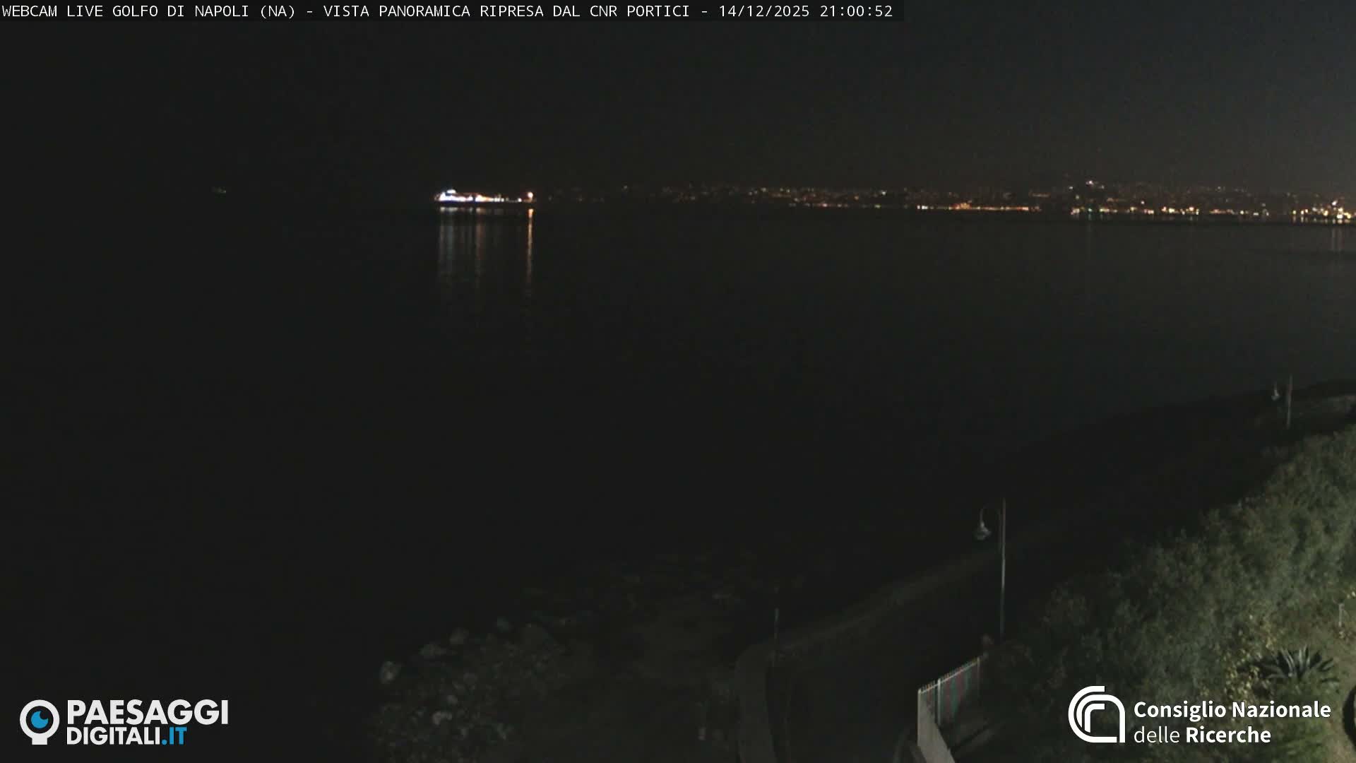 A panoramic view captures a calm bay with a rocky shoreline and a paved promenade on the right, leading to a distant city and mountains under a bright, partly cloudy sky, with a few boats visible on the water.