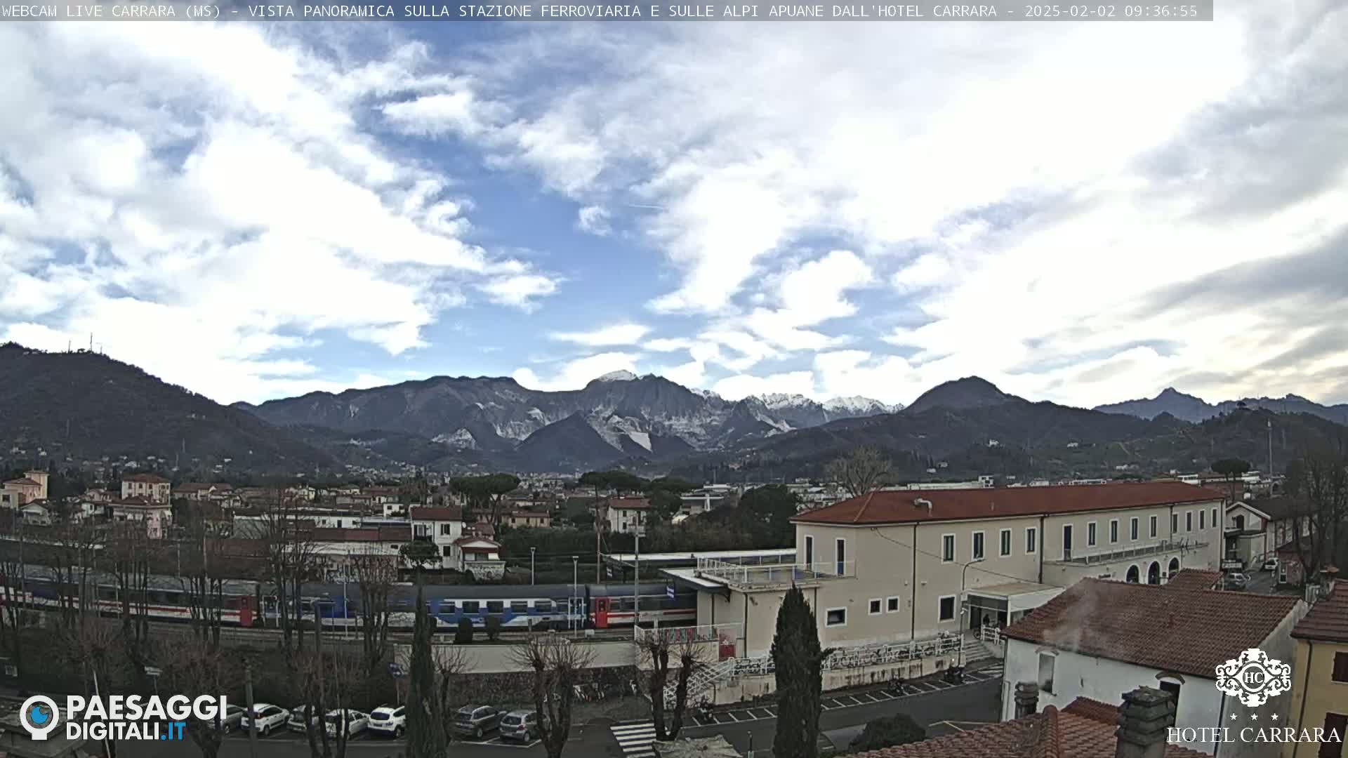 A partly cloudy day reveals a train at a station in a town nestled in a valley, with snow-capped mountains in the background.