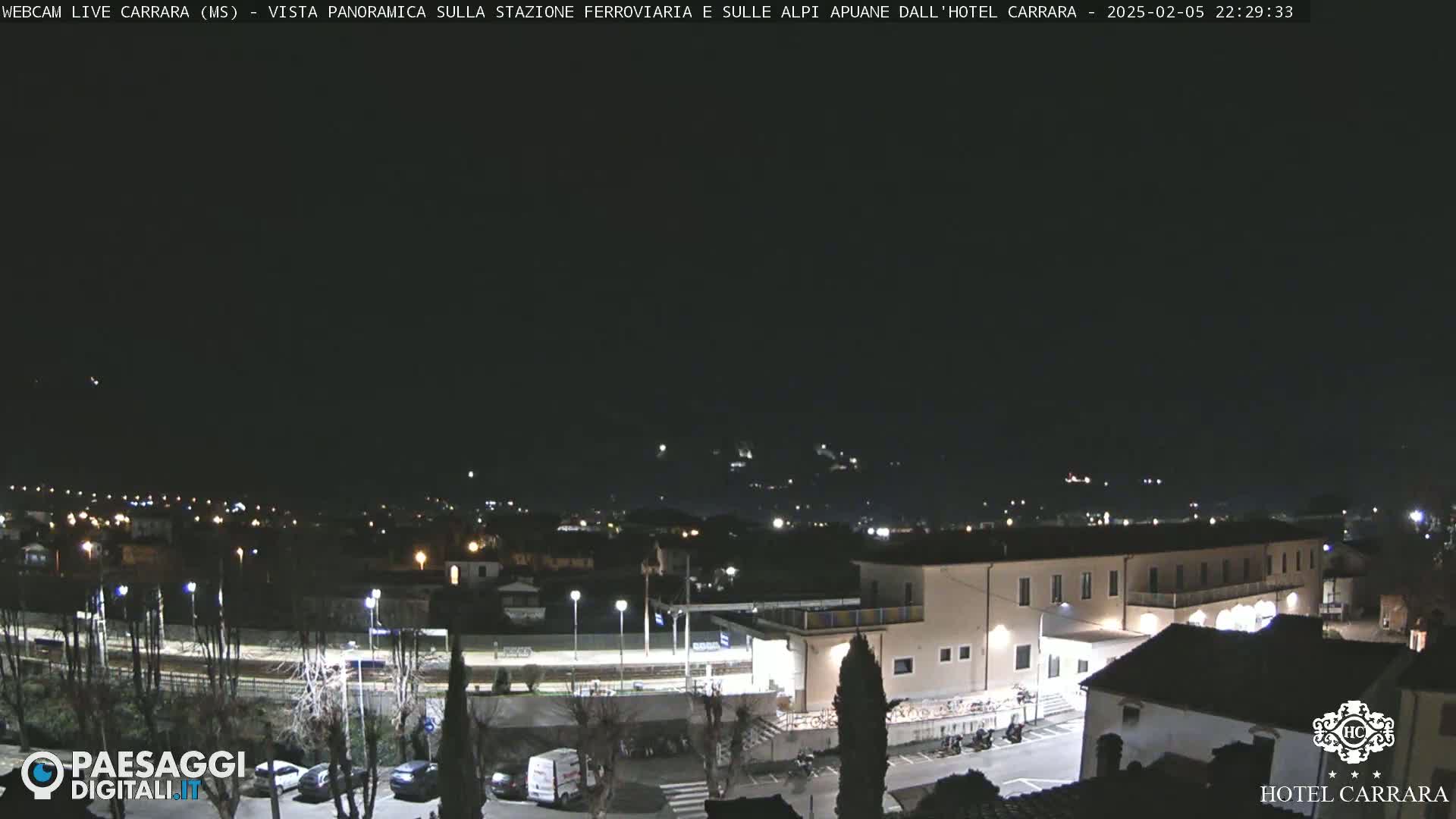 A nighttime, panoramic view of a town's outskirts, including a train station and a building, under a clear, dark sky.