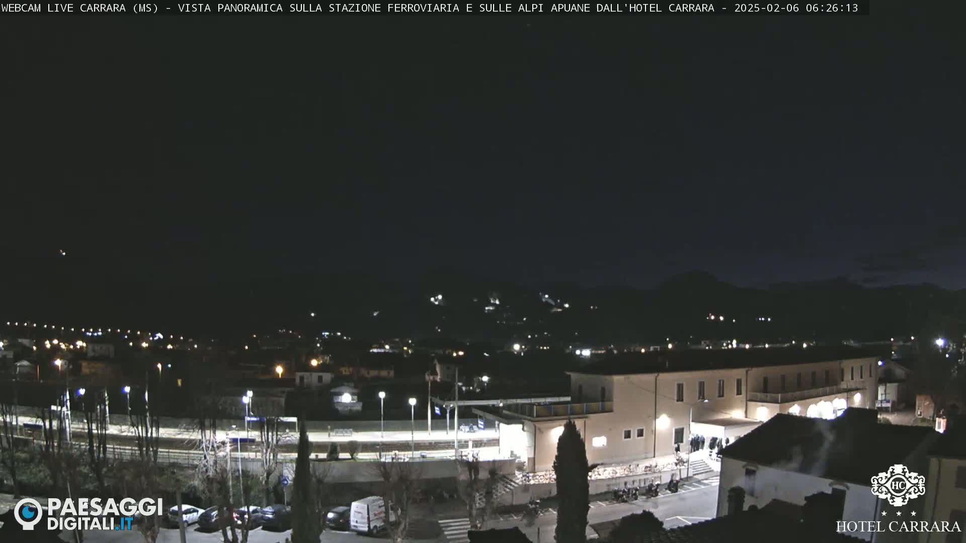A nighttime, panoramic view of a town's train station and surrounding buildings, with hills in the background under a dark sky.