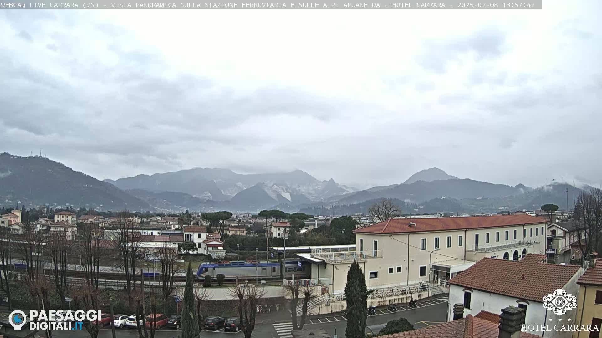 A train sits at a station in a town nestled in a valley, with overcast skies and mountains in the background.