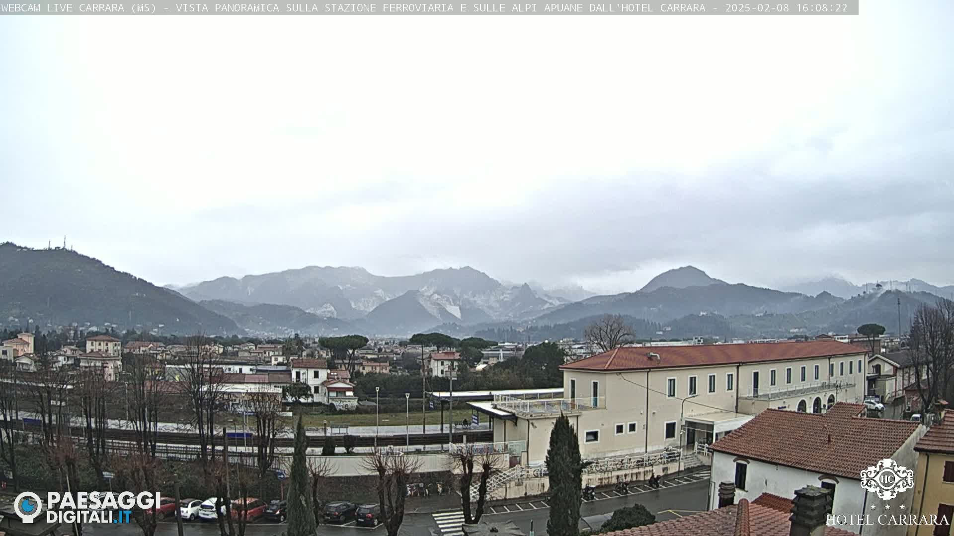 A long shot of a town nestled in a valley, under an overcast sky, with snow-capped mountains in the background.