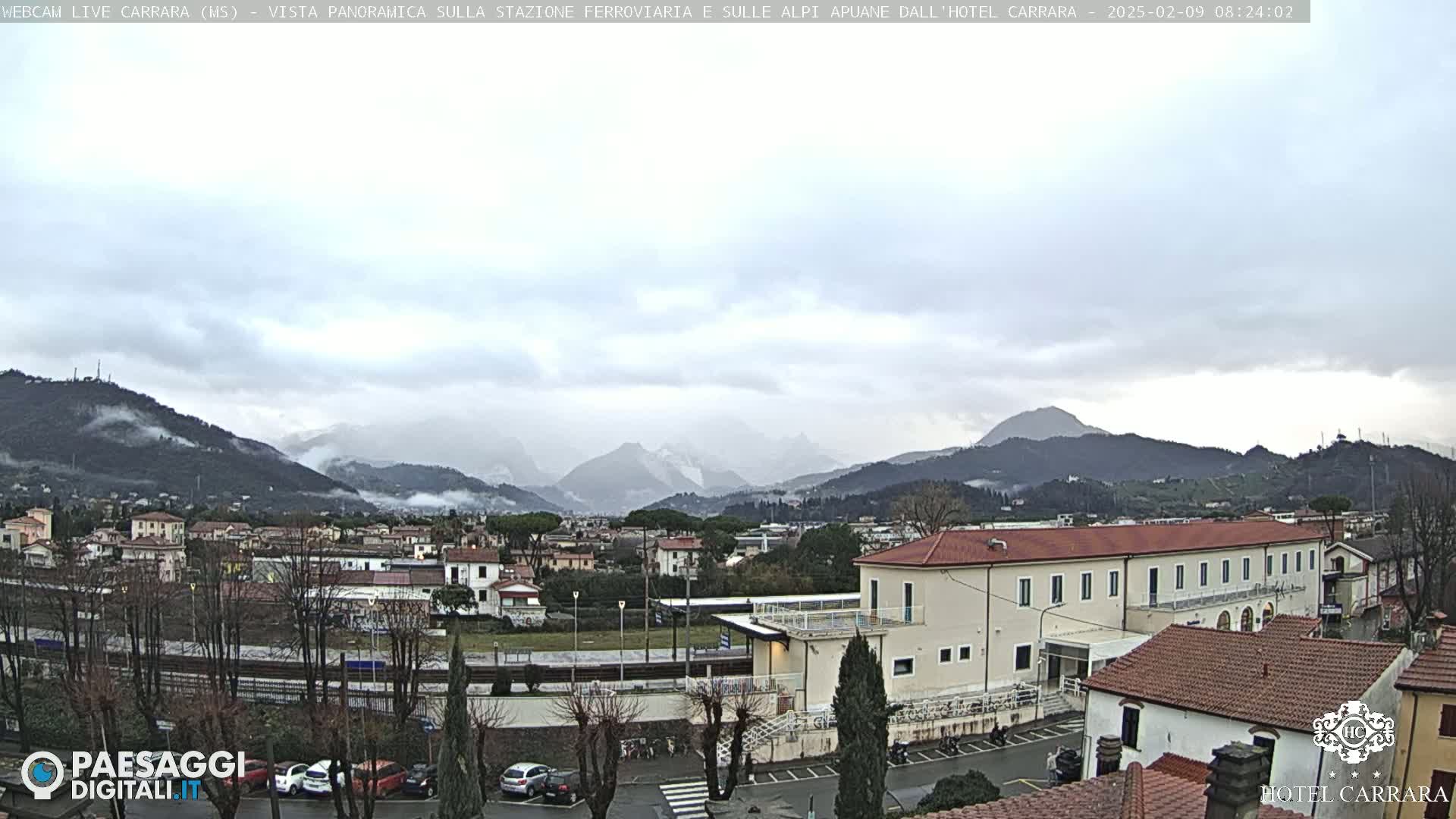 A panoramic view of a town nestled in a valley, with snow-capped mountains in the background under a cloudy sky.